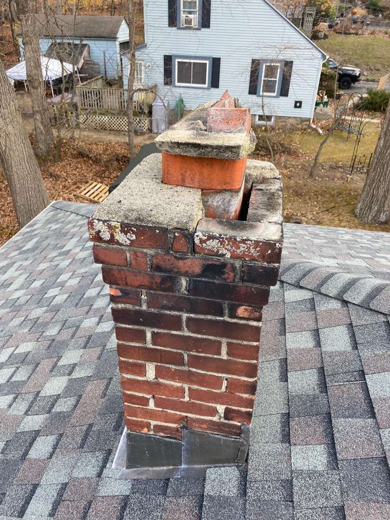 Damaged brick chimney on a gray shingled roof with missing bricks and a broken cap