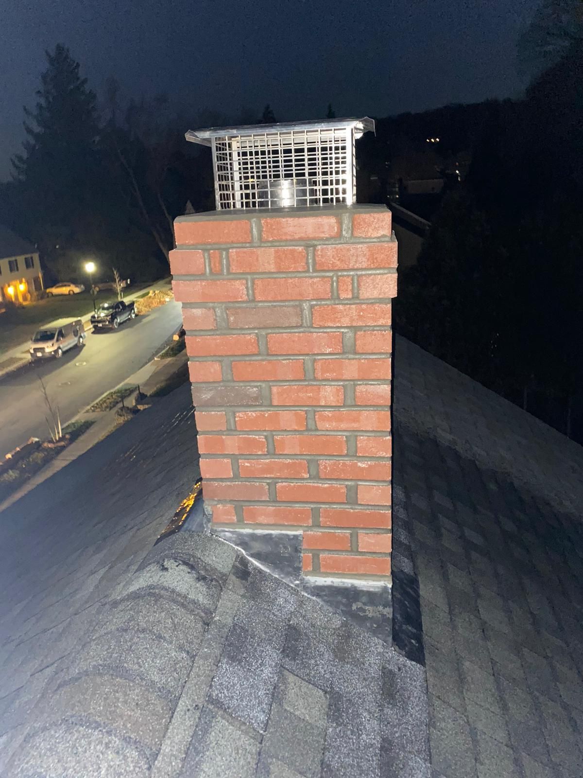 Brick chimney on a shingled roof at night, with a metal cap and a street visible below.