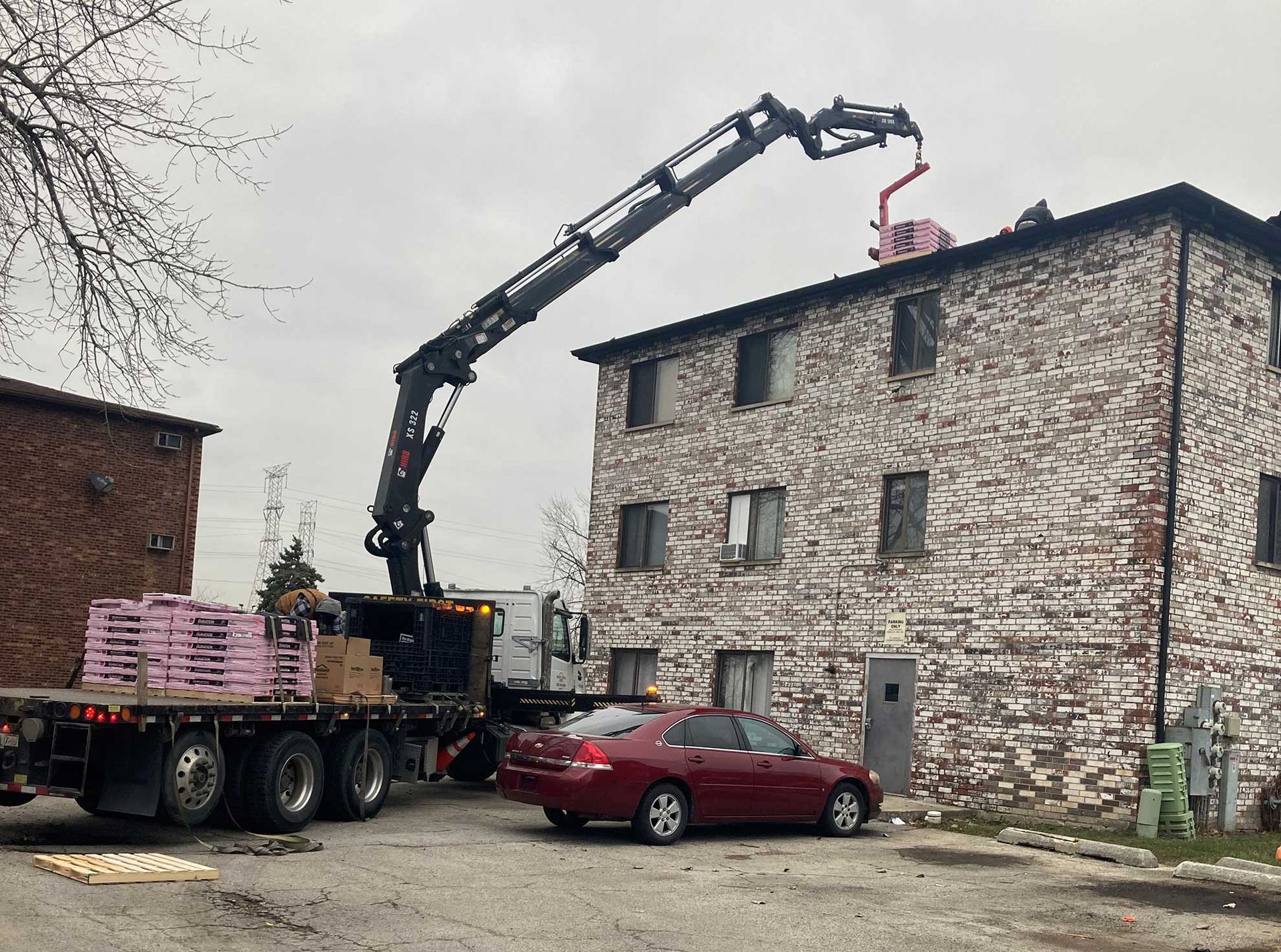 A crane lifts materials onto the roof of a brick apartment building. A flatbed truck holds building supplies.