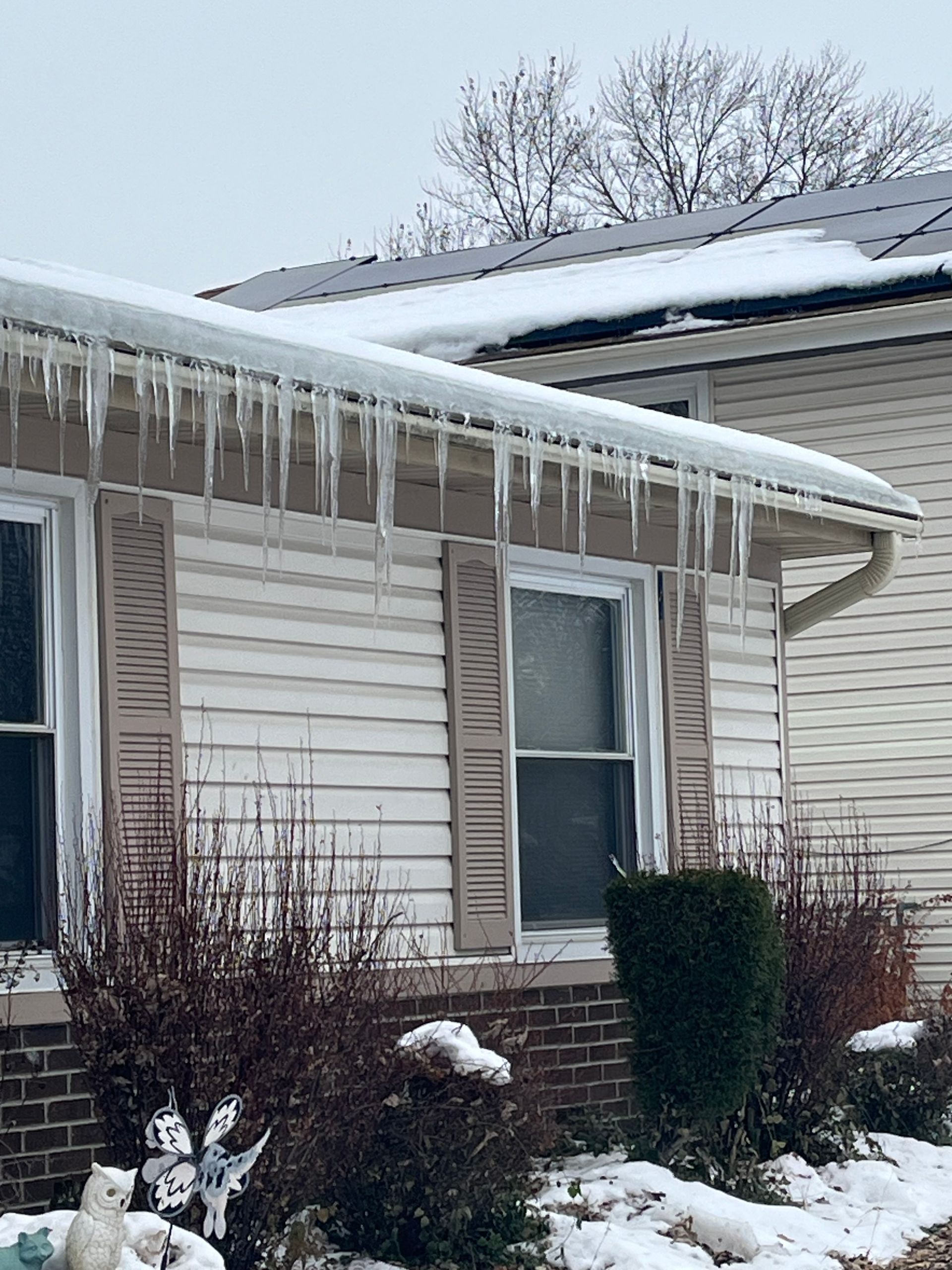 Icicles hanging from a house's roofline. Snow on the ground and roof. Shutters on windows.