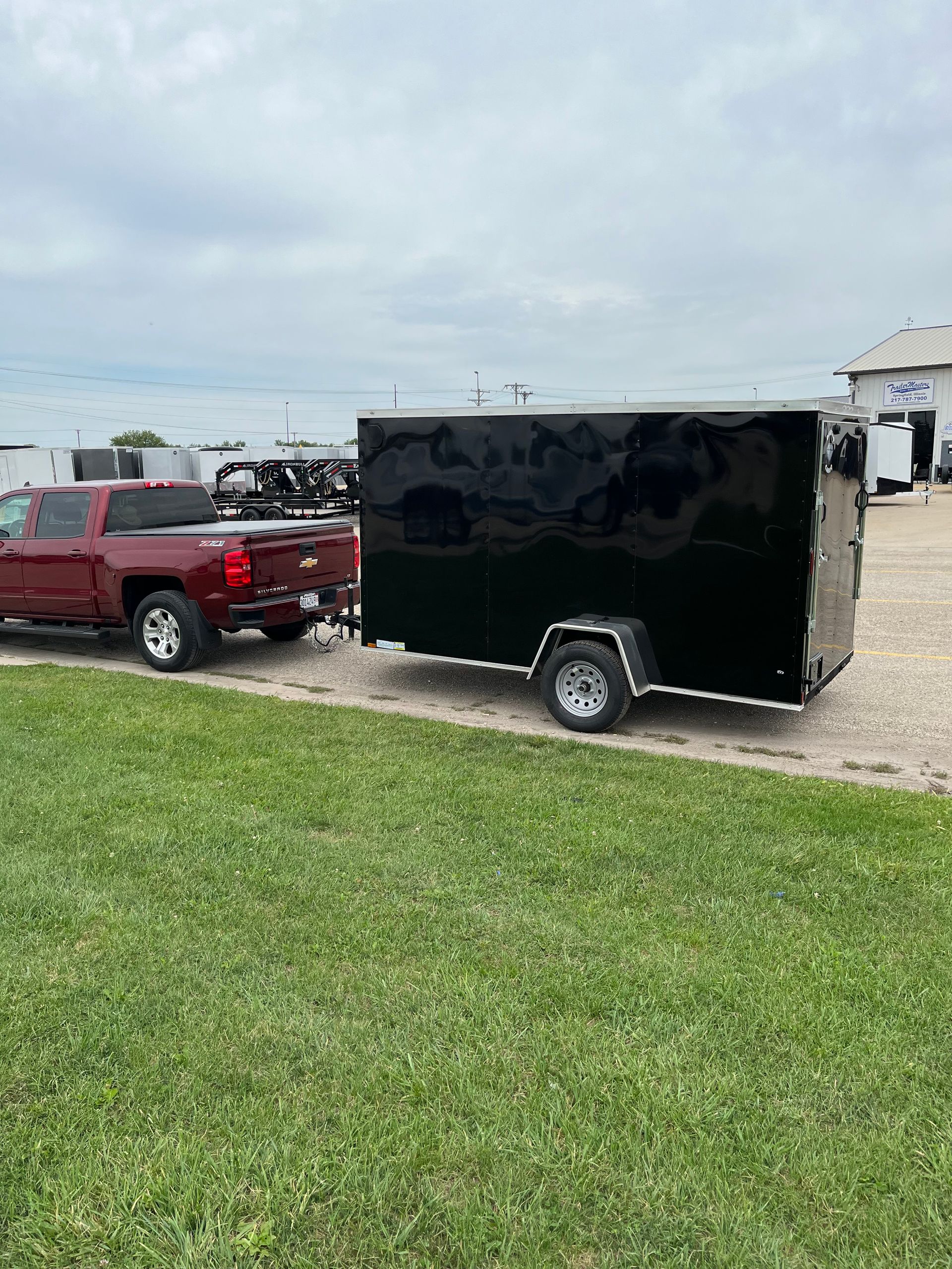 Red pickup truck towing a black enclosed trailer on a gravel lot near grass. Cloudy sky.