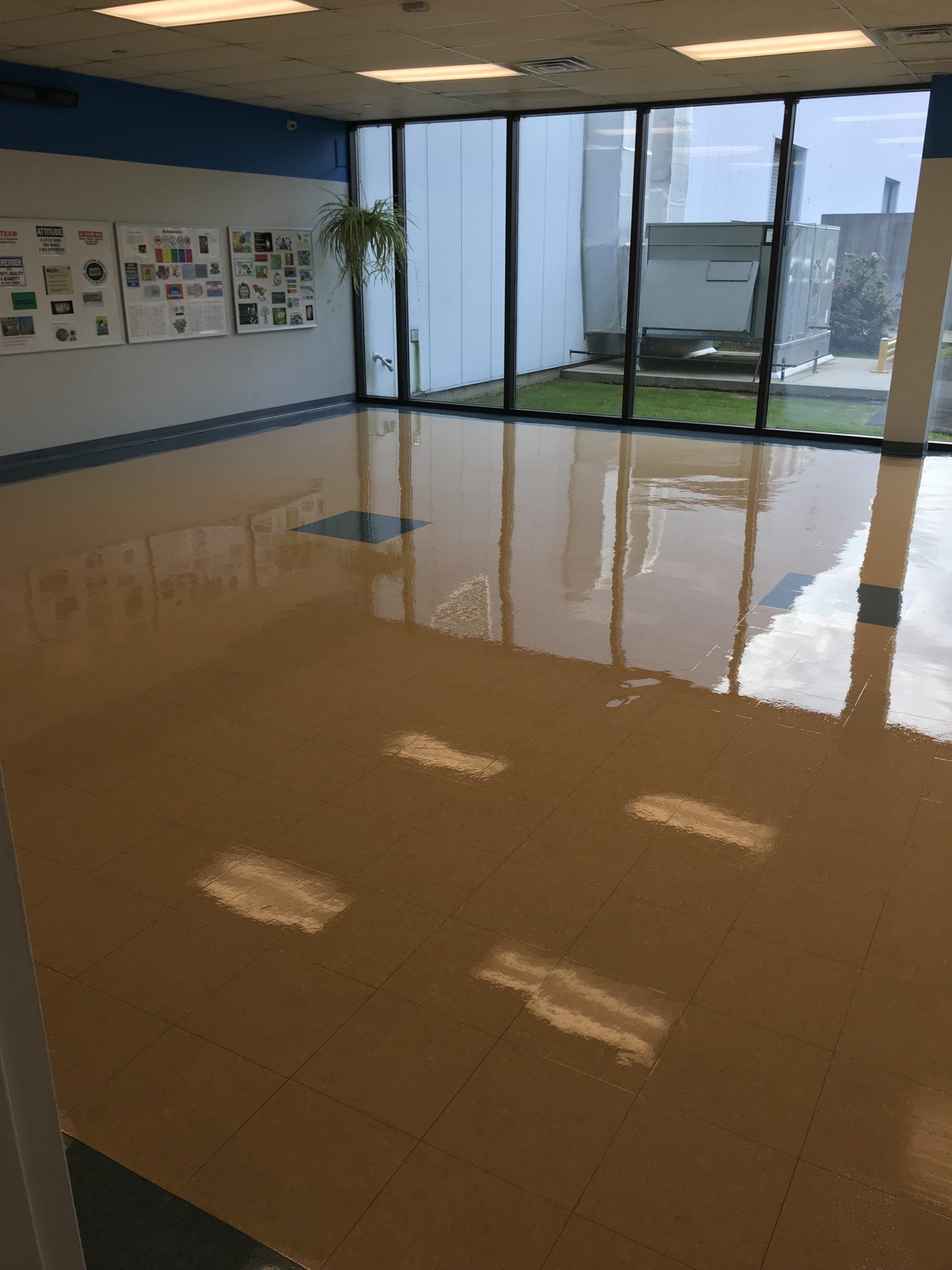 A room with a shiny, light-colored floor, a wall with papers, and large windows looking onto an outdoor area.