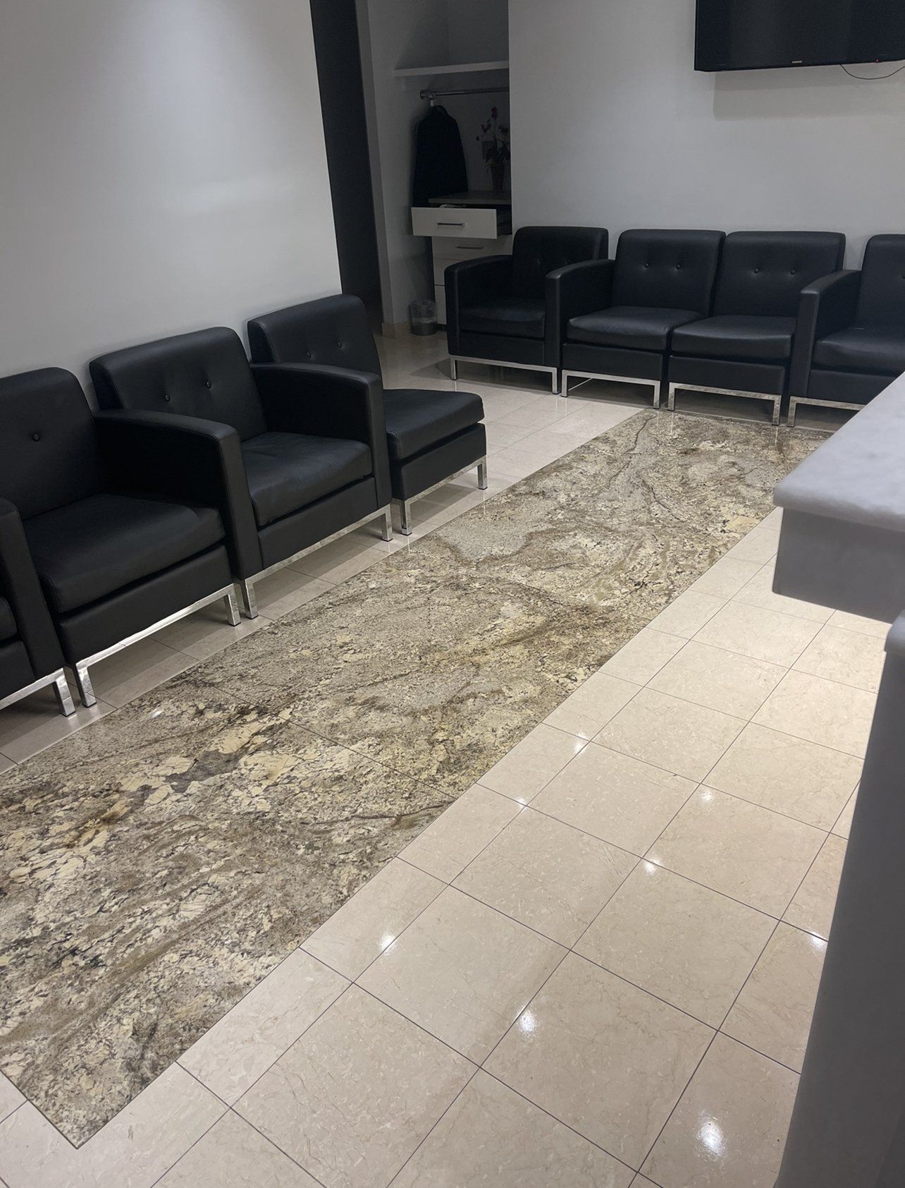 A waiting room with black leather chairs arranged along a patterned rug, tile floor. A coat rack in background.