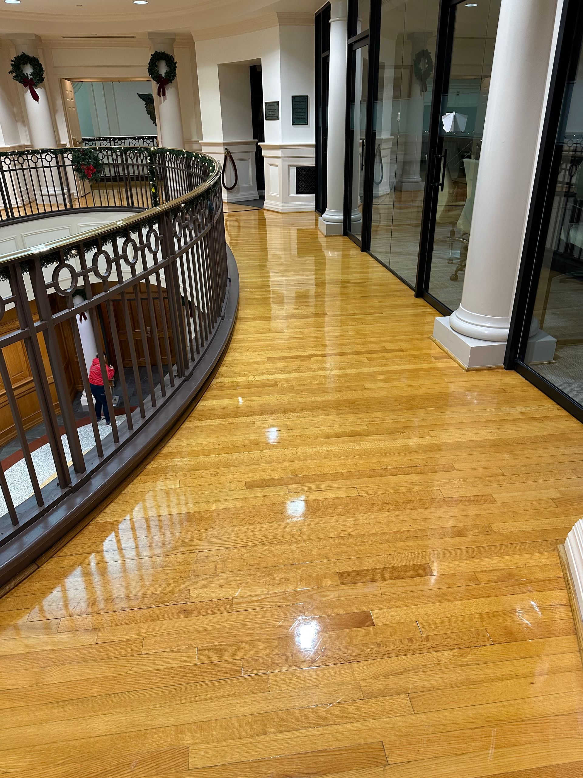 Shiny, wooden floor curves through a building corridor. Wrought iron railing, and windows and pillars in background.