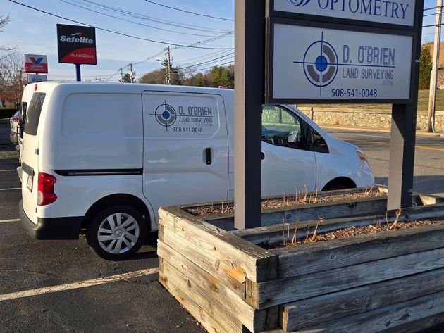 A white O'Brien Land Surveying van parked next to a matching sign in a parking lot.
