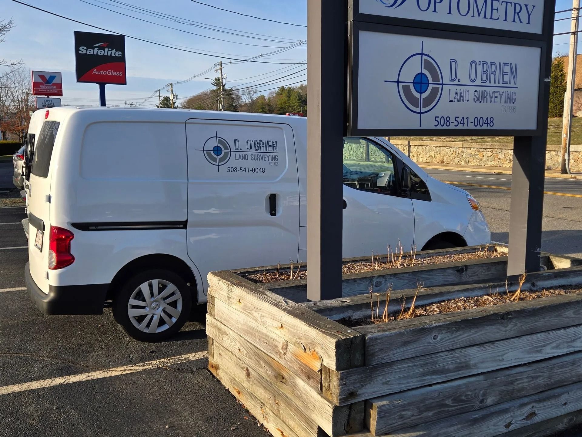 A white work van parked next to a wooden planter and an optometry business sign.