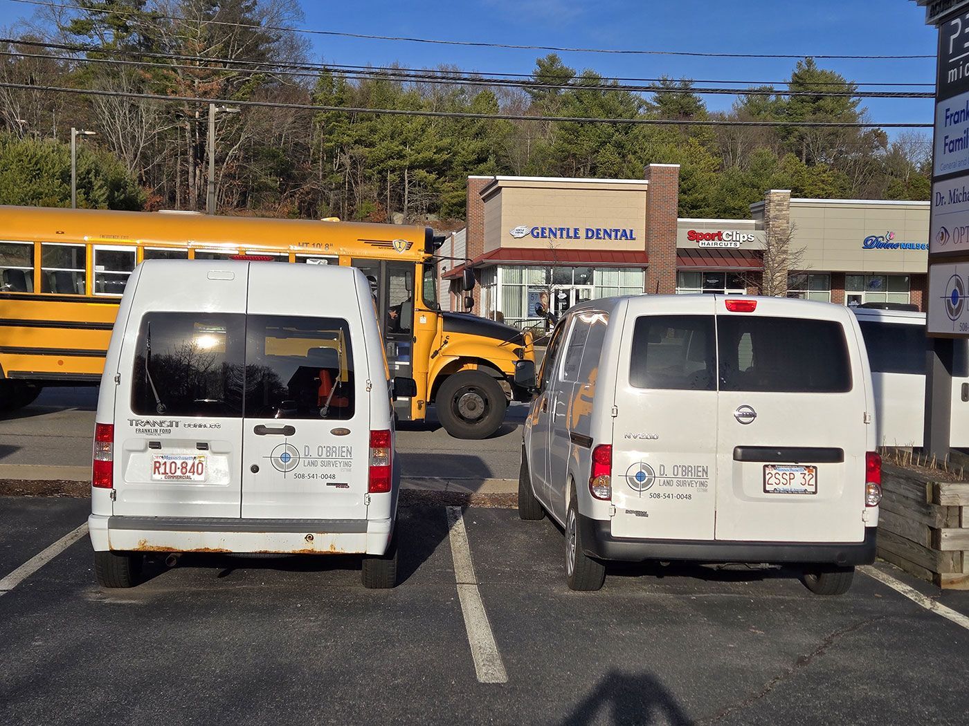 Two white cargo vans parked in a lot in front of a shopping center with a yellow school bus in the background.