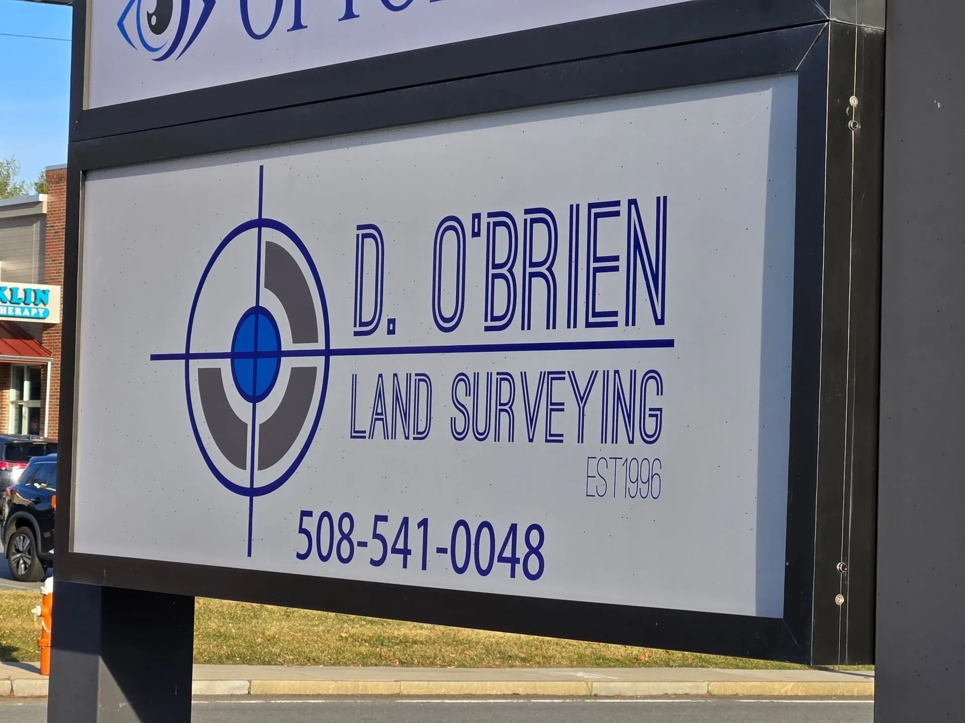 A white sign for D. O’Brien Land Surveying with a blue and gray target logo, the phone number 508-541-0048, and "est. 09."