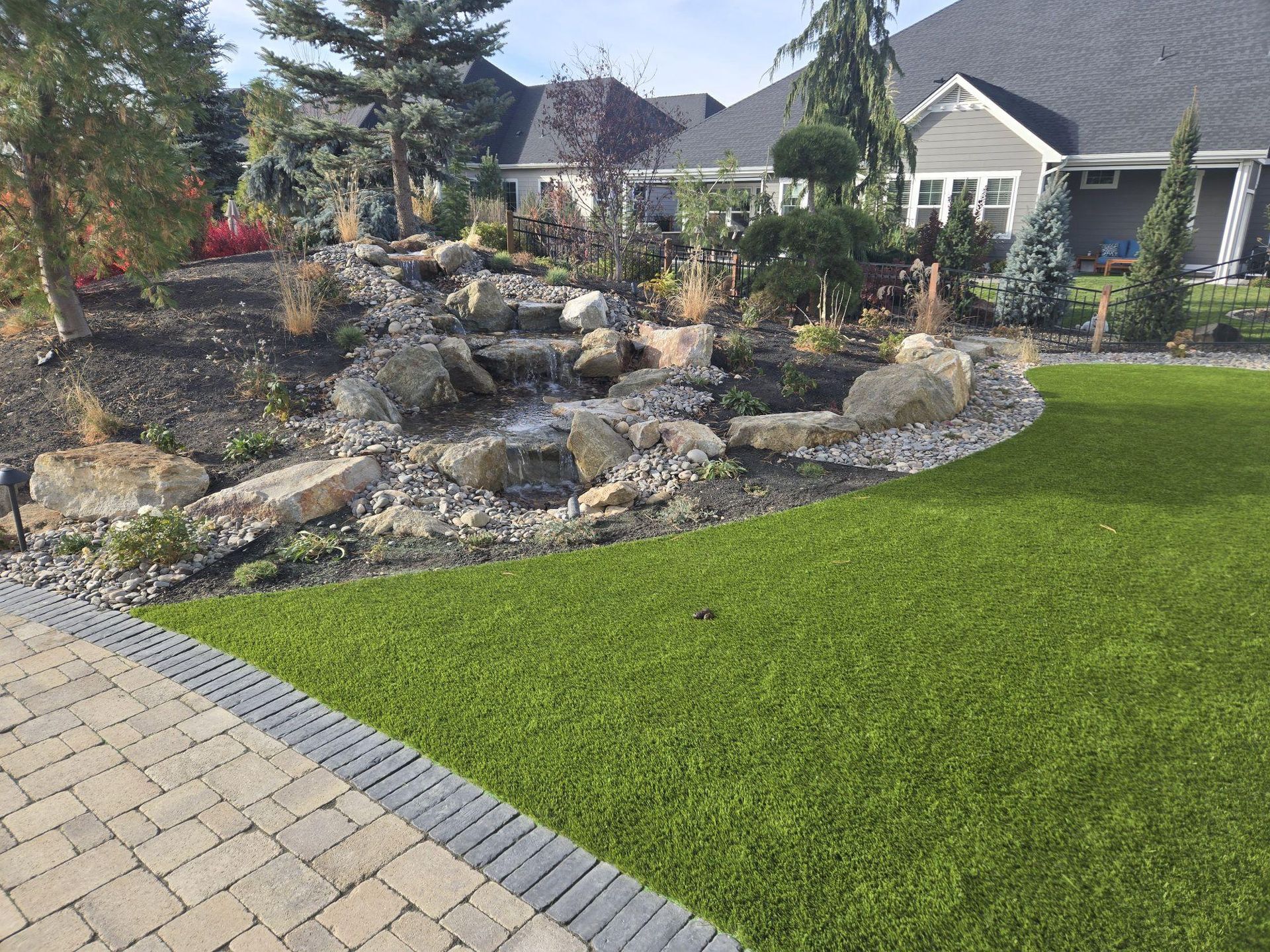 Landscaped backyard with a rock waterfall, green lawn, and stone patio beside a house