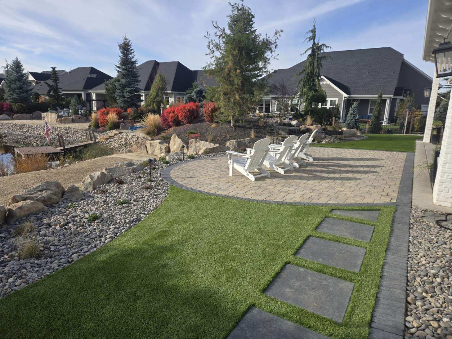 Landscaped backyard patio with white chairs, stone path, grass lawn, and modern houses in the background