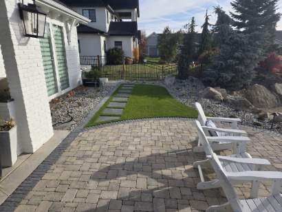 Backyard patio with stone pavers, green lawn strip, and white chairs beside a house