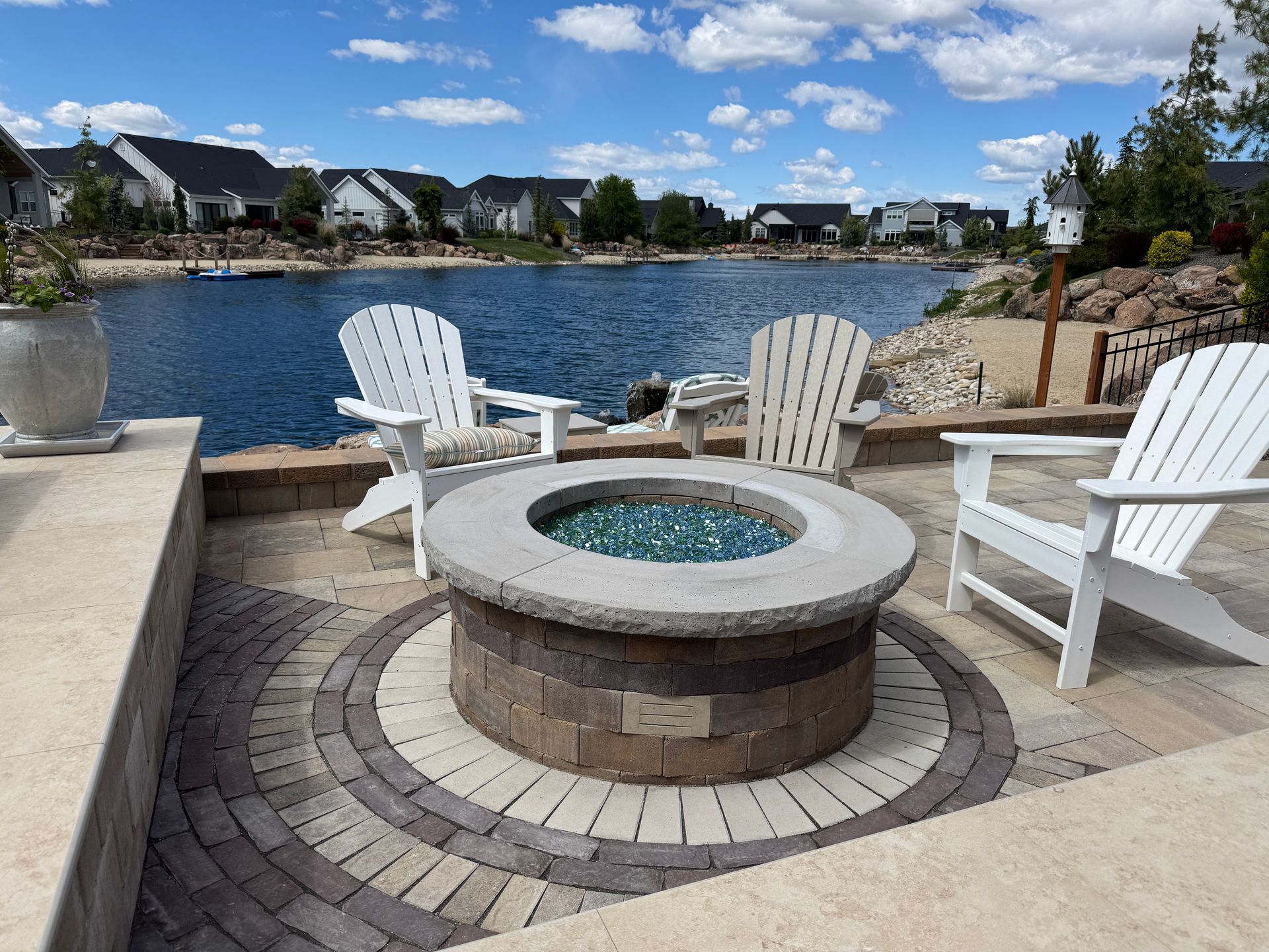 Lakeside patio with white chairs вокруг круглого stone fire pit under a blue sky
