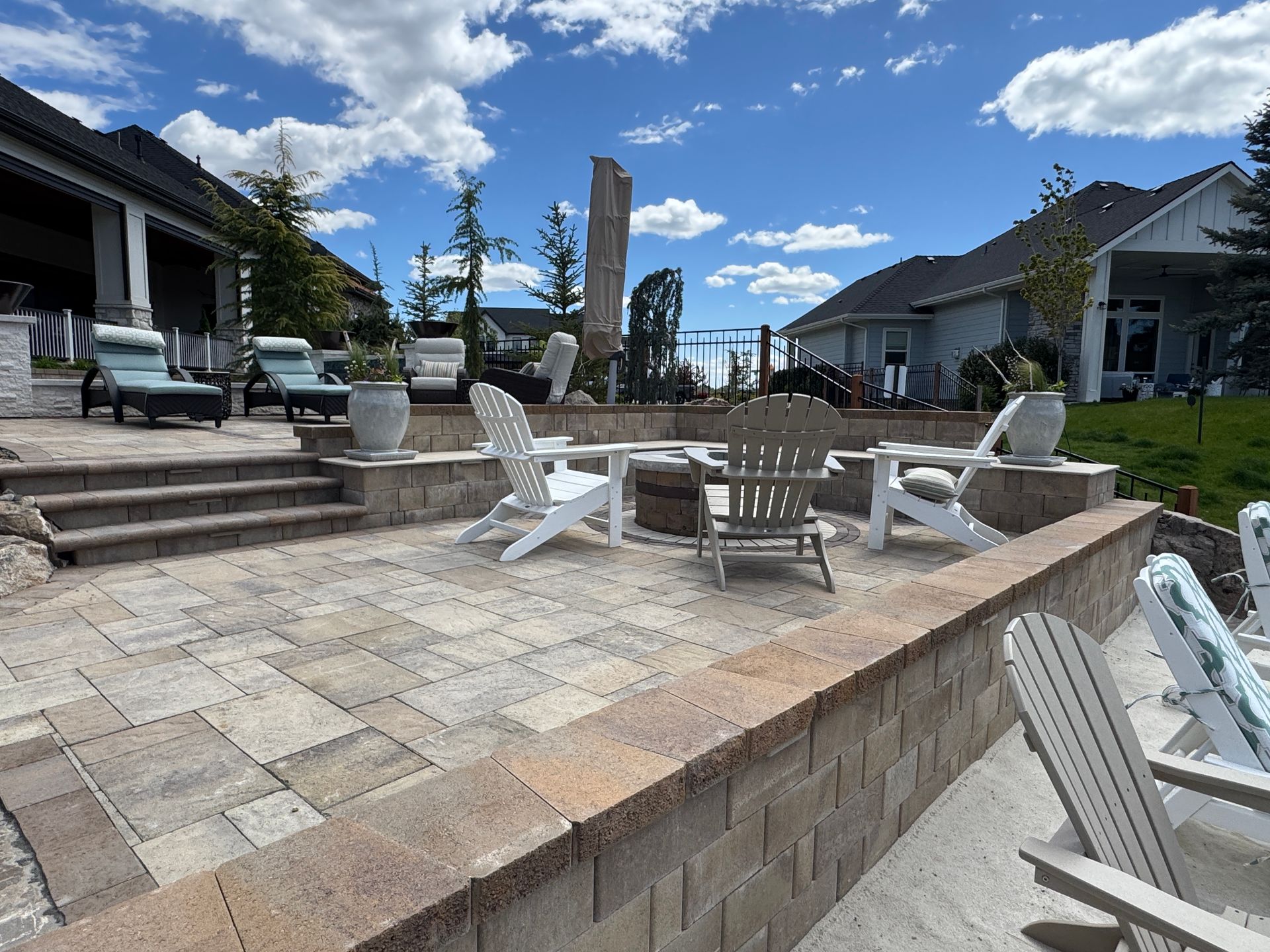 Patio with white lounge chairs and fire pit surrounded by houses under a blue sky with clouds
