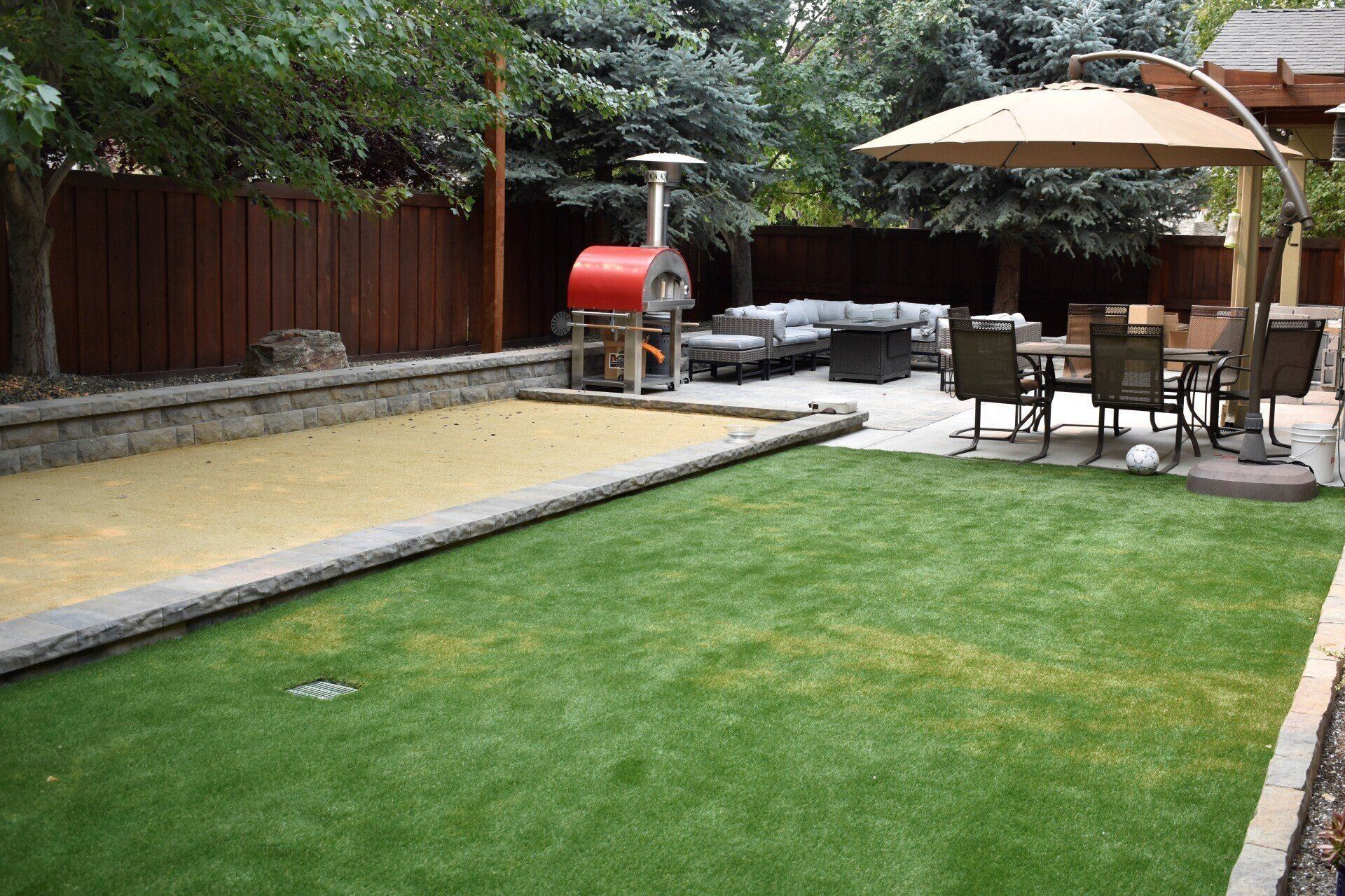 Backyard with green lawn, stone patio, outdoor dining set, grill, and umbrella under trees