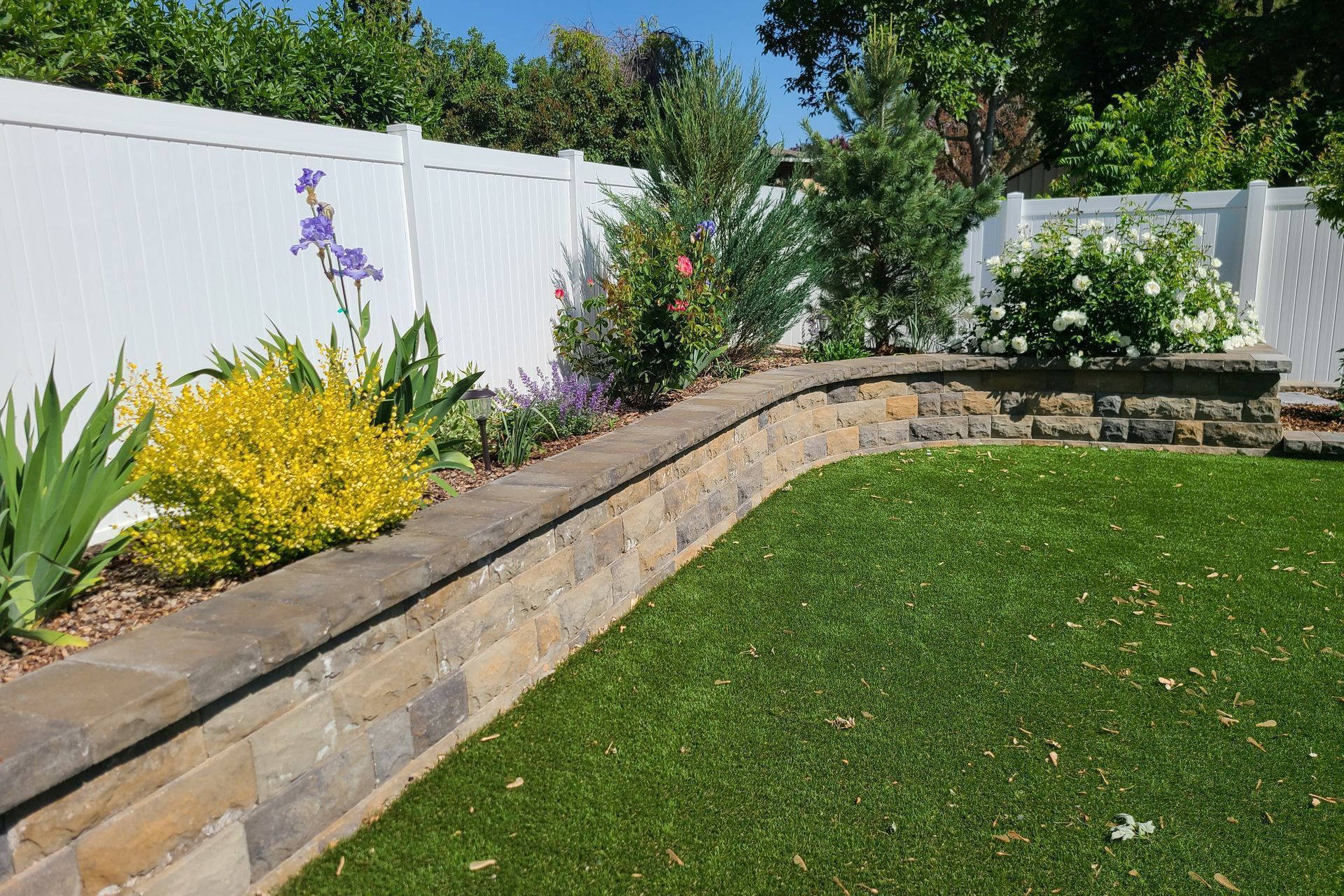 Curved stone garden bed with colorful flowers and shrubs beside a green lawn and white fence