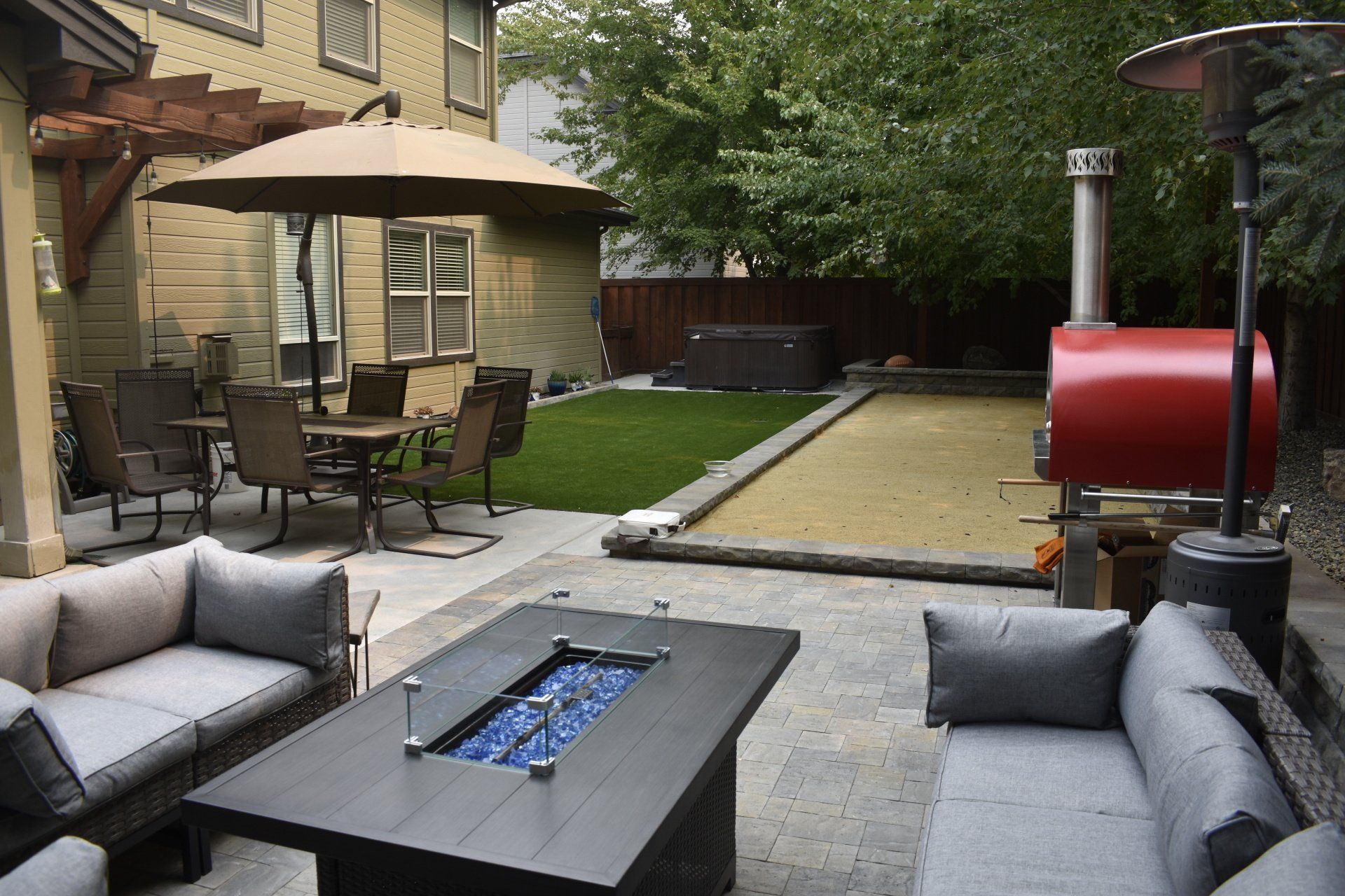Backyard patio with gray seating, fire pit, dining table, and covered grill beside a putting green