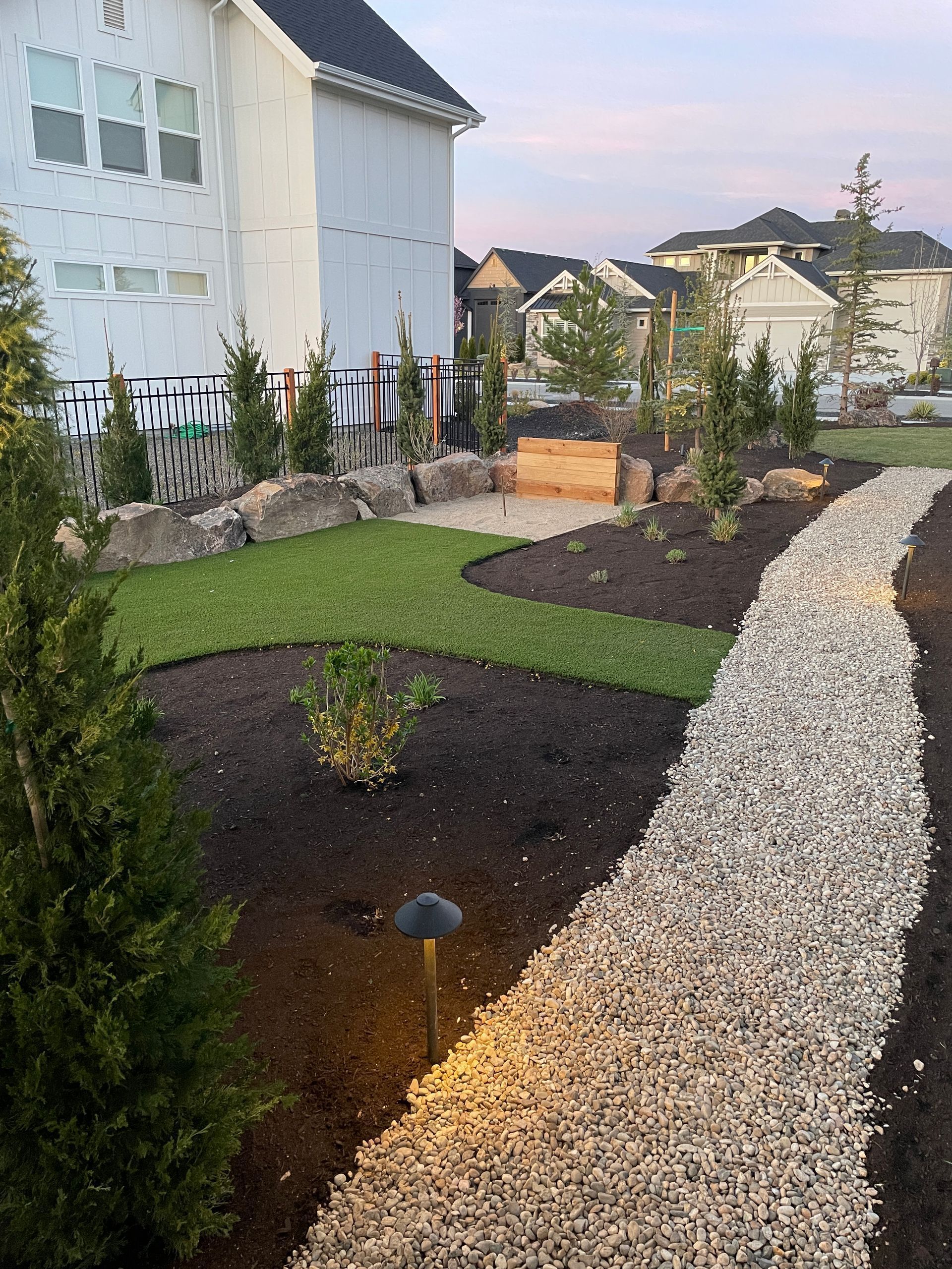 Landscaped yard with gravel path, dark mulch beds, shrubs, and houses in the background at dusk