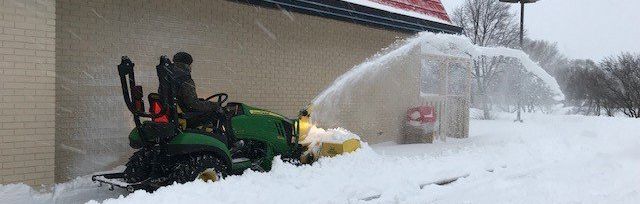 A green tractor snowblower clears snow in front of a building