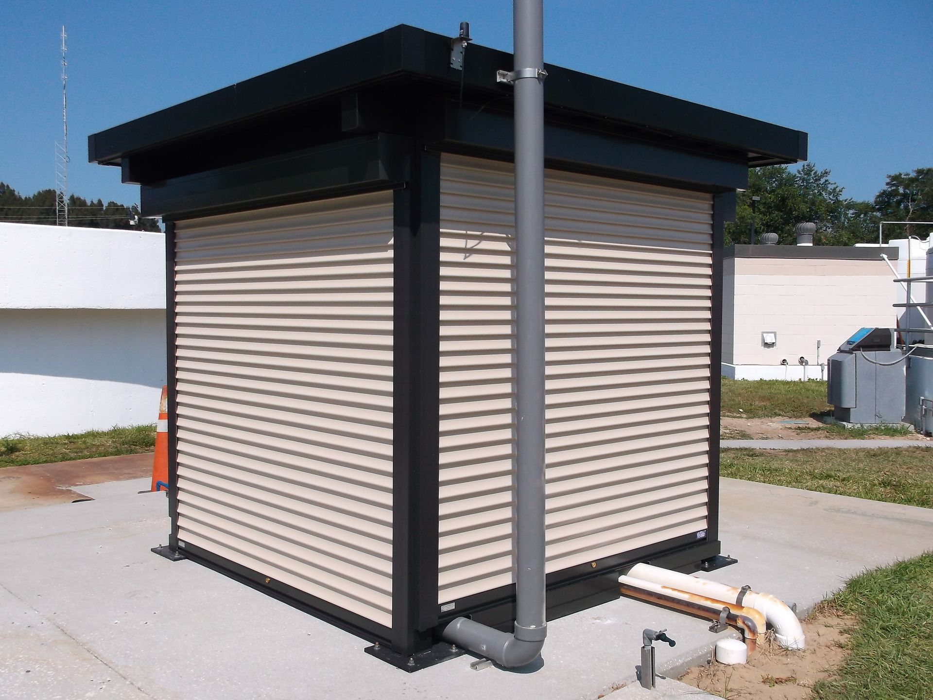 Small beige utility shed with black trim and a sloped roof beside a metal pole outdoors.