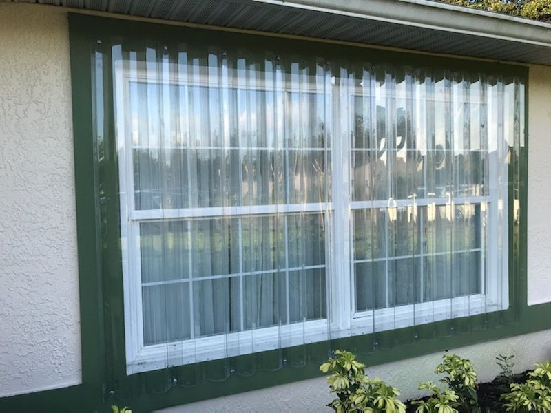 White window with sheer curtains on a green house exterior, with a small bush below