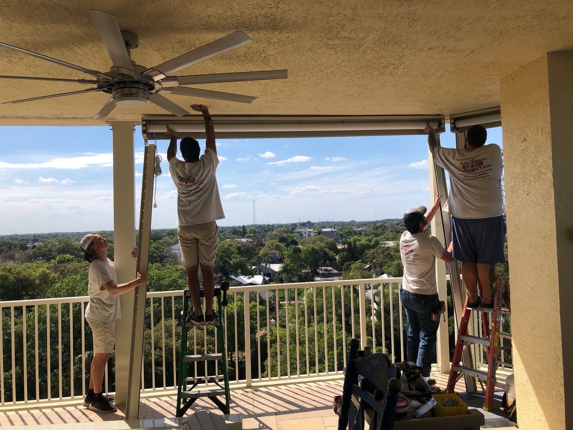 Four people stand on ladders fixing a ceiling fan on a balcony overlooking a city.
