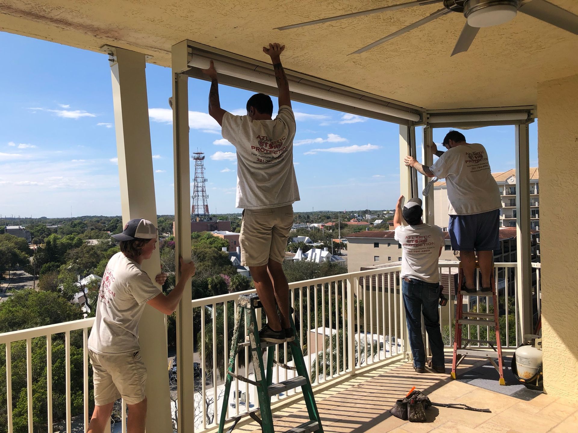Workers installing ceiling panels on a balcony, with ladders, tools, and a city view beyond the railing