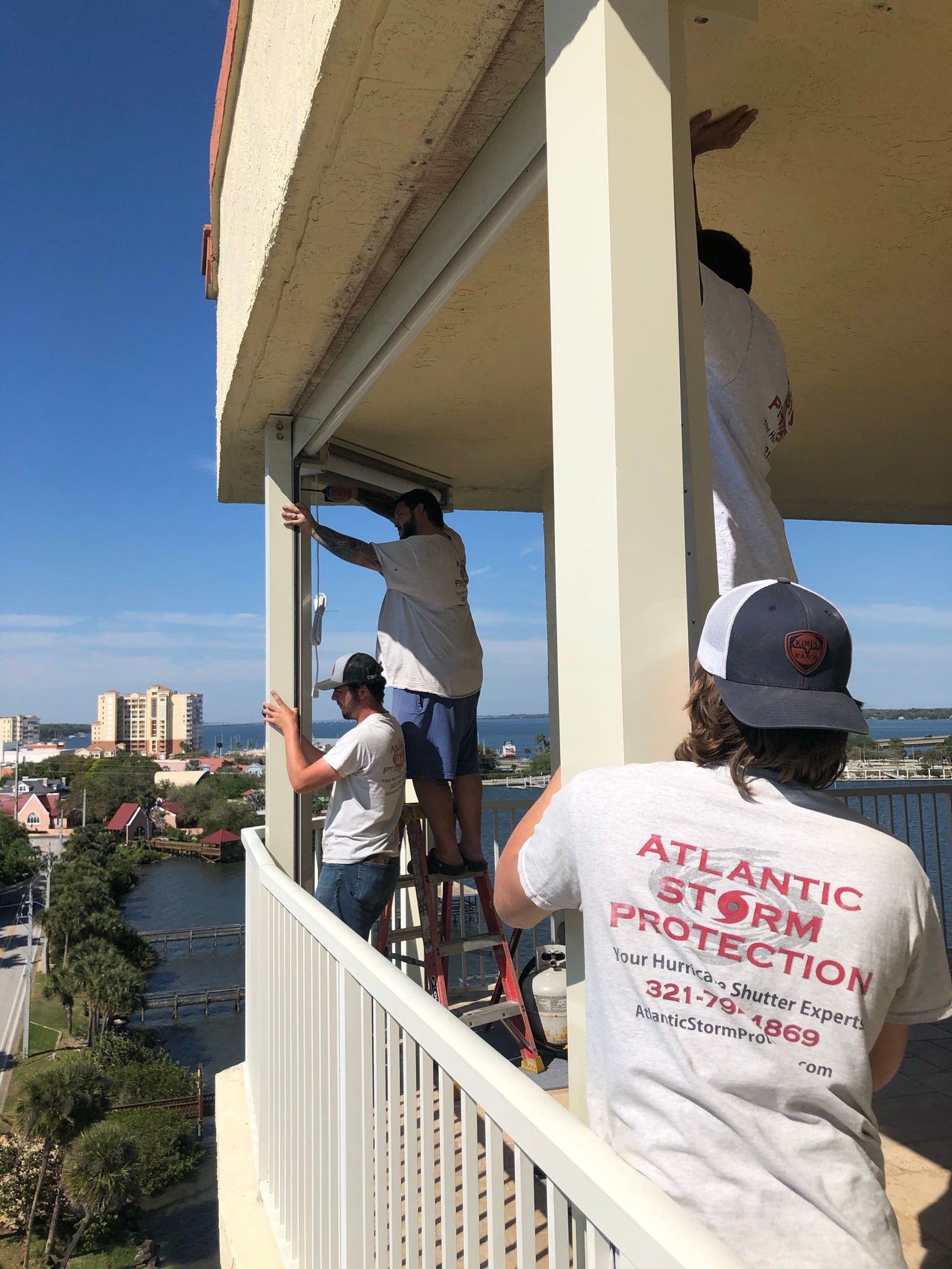 Workers painting a high-rise balcony railing, one on a ladder, with city view beyond