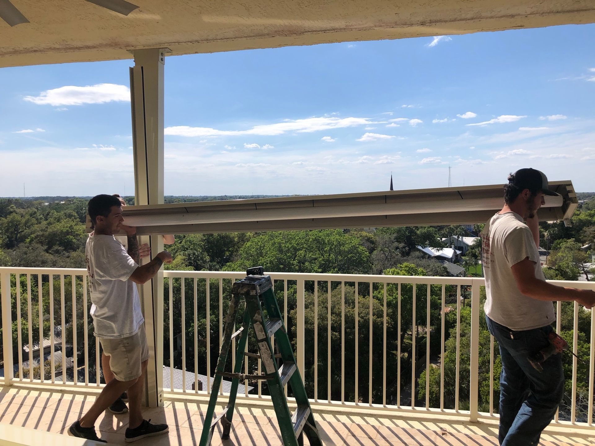 Two workers move a long beam through a covered outdoor area with a tripod on the floor.