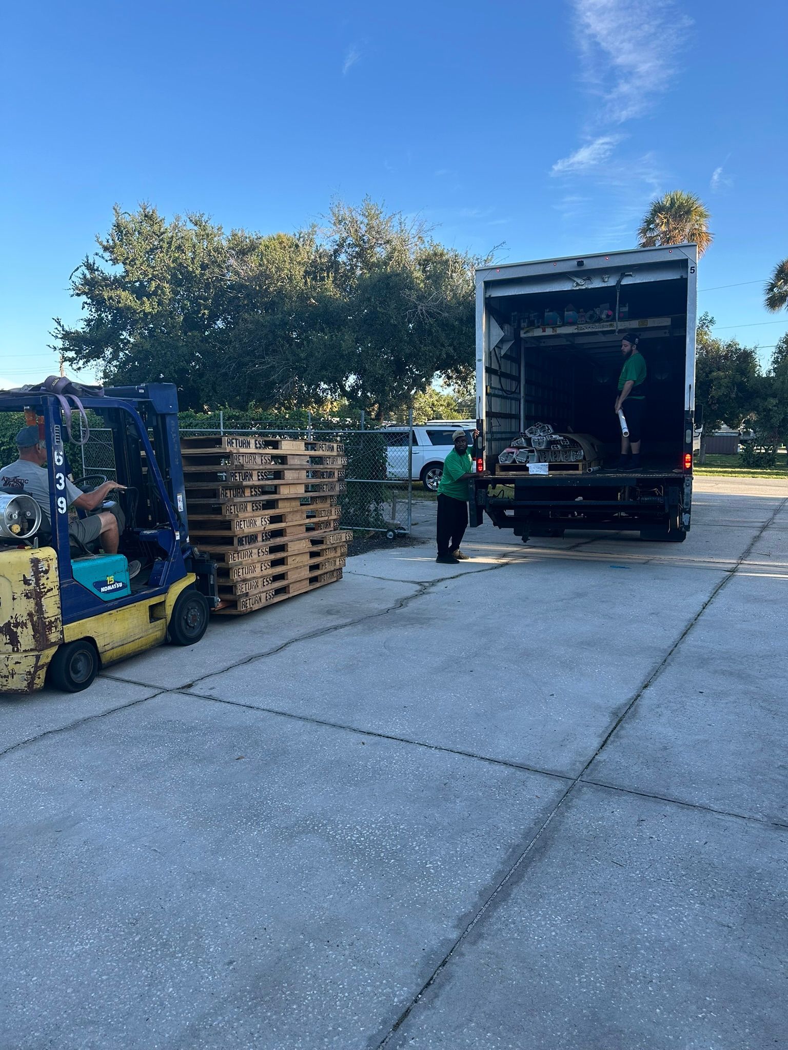 Forklift and truck loading wooden pallets on a concrete lot under a blue sky