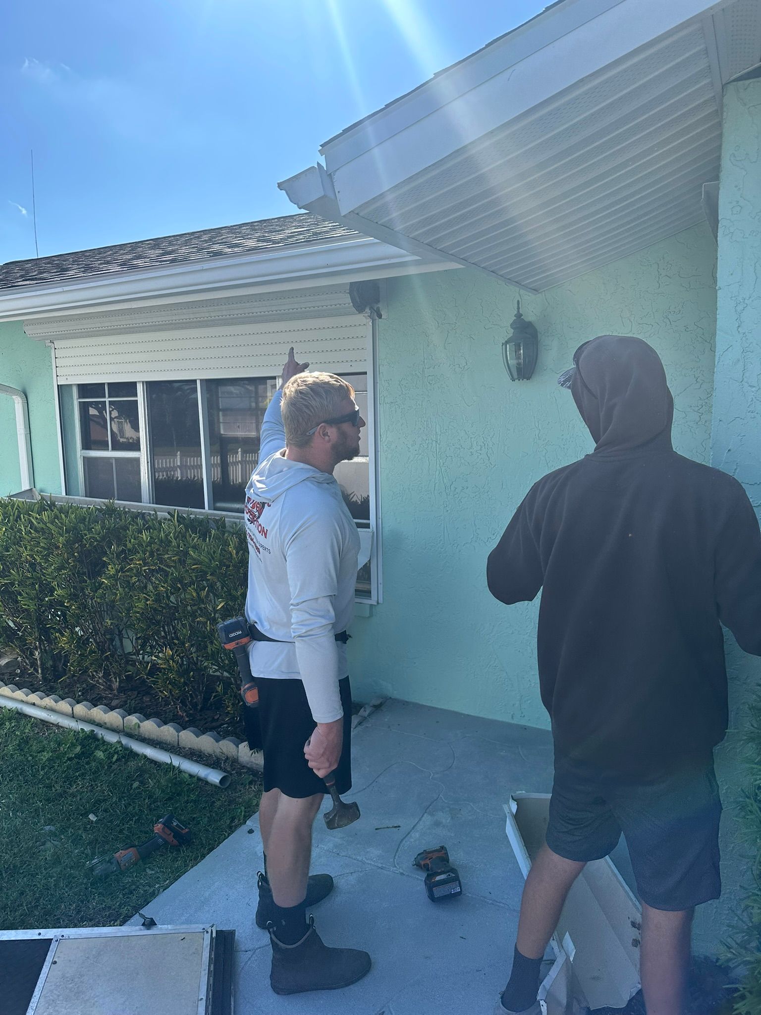 Two people stand by a turquoise house with hedge and windows, one holding a bag and the other facing him.