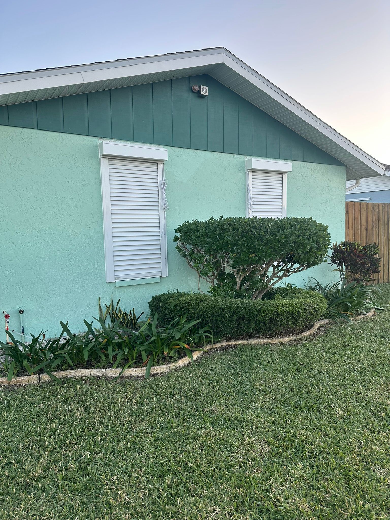 Mint-green house exterior with white shutters and a trimmed shrub in front of a lawn