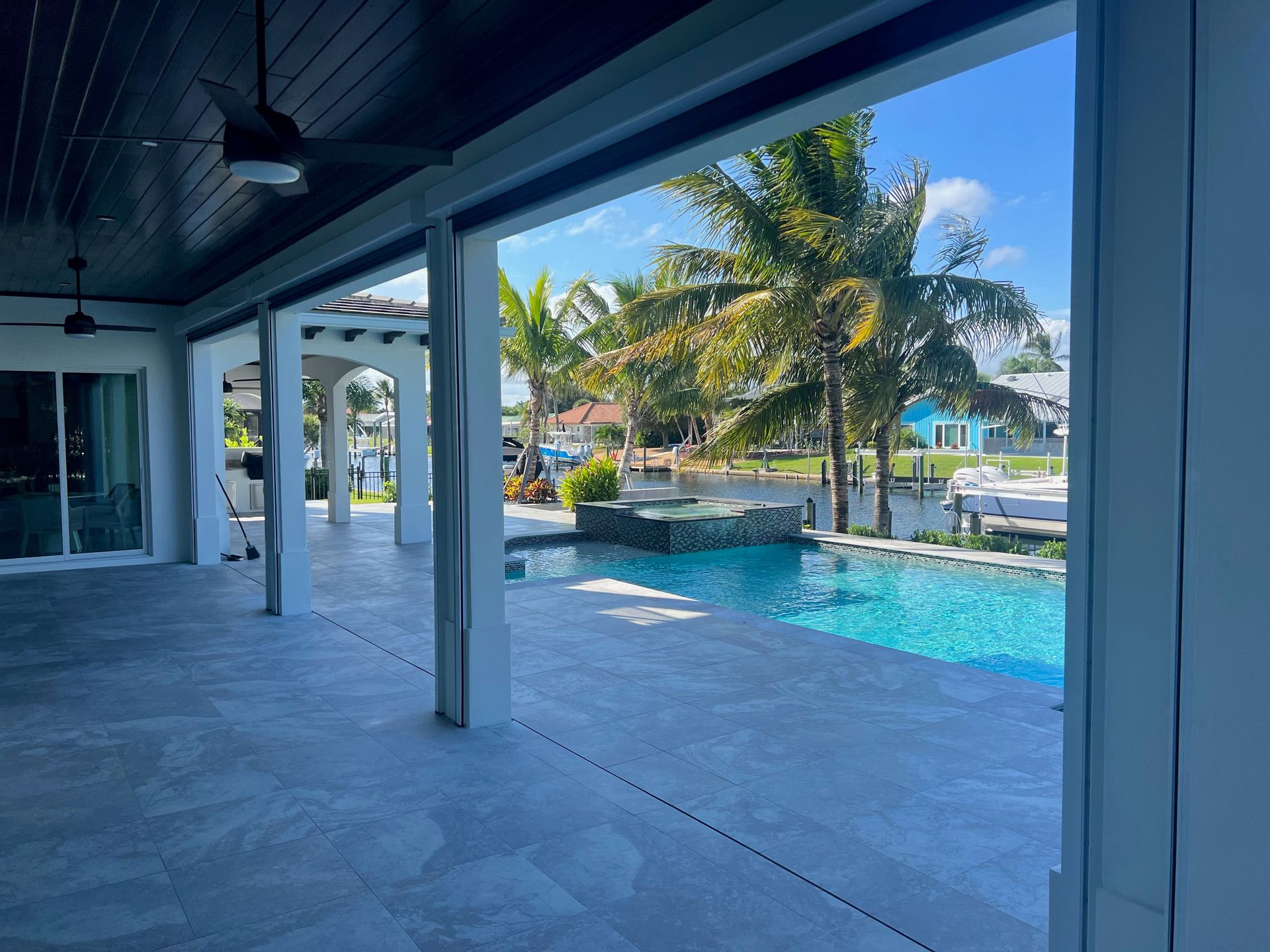 Covered patio overlooking a turquoise pool, palm trees, and a sunny tropical waterfront view.