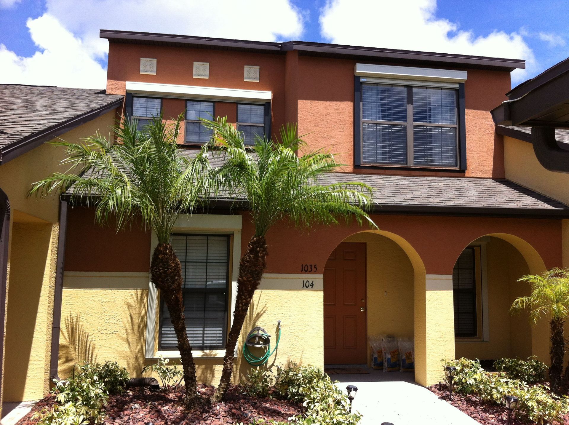 Two-story tan house with brown trim, arched entryway, and palm trees in front under a blue sky