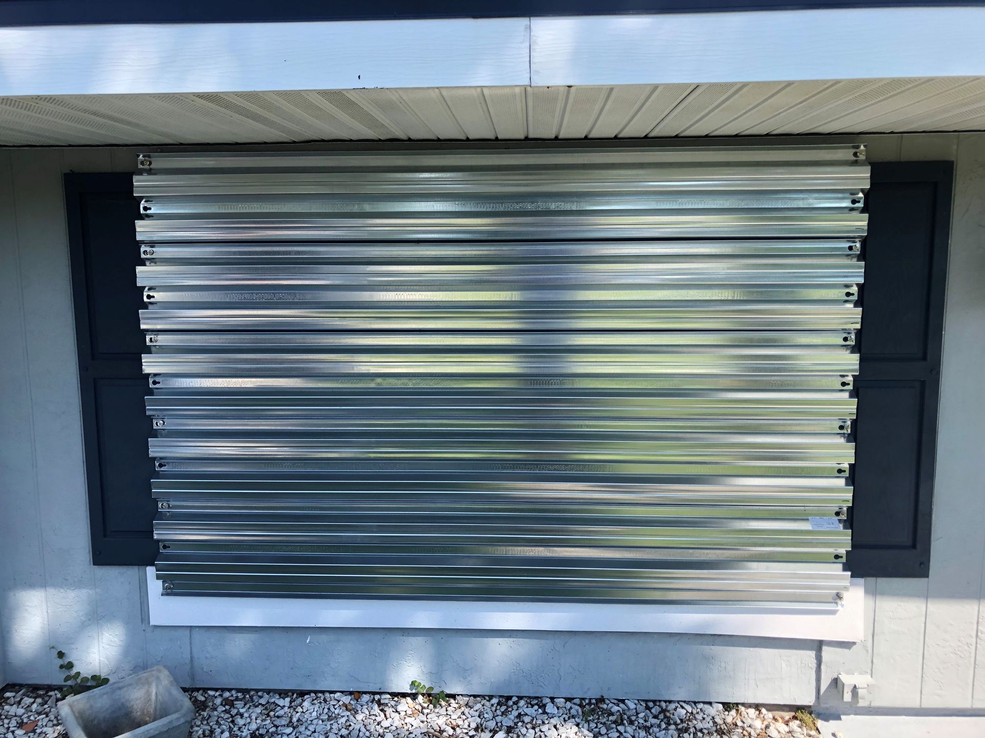 Shuttered front window on a white house, covered with a metal security barrier and flanked by black shutters