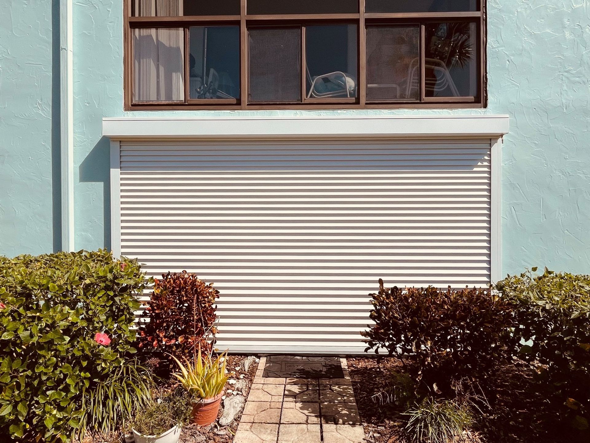Closed white shutter beneath a window on a pastel blue building, framed by bushes and a stone path