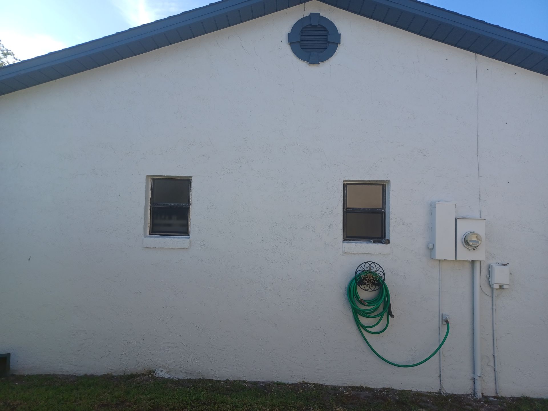 White building exterior with two small windows, a round vent, and a green garden hose on the wall.