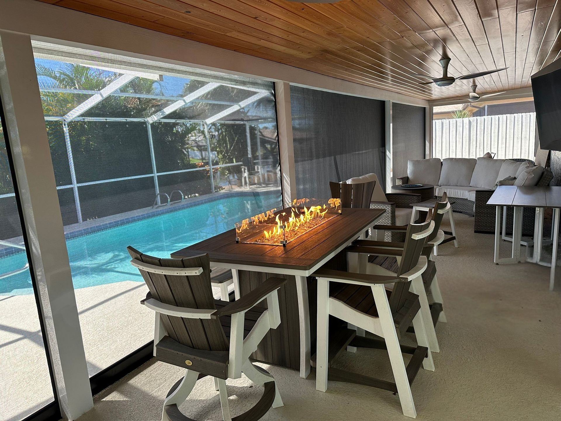 Covered patio dining area beside a pool, with a long wooden table, chairs, and warm candlelight under a wood ceiling