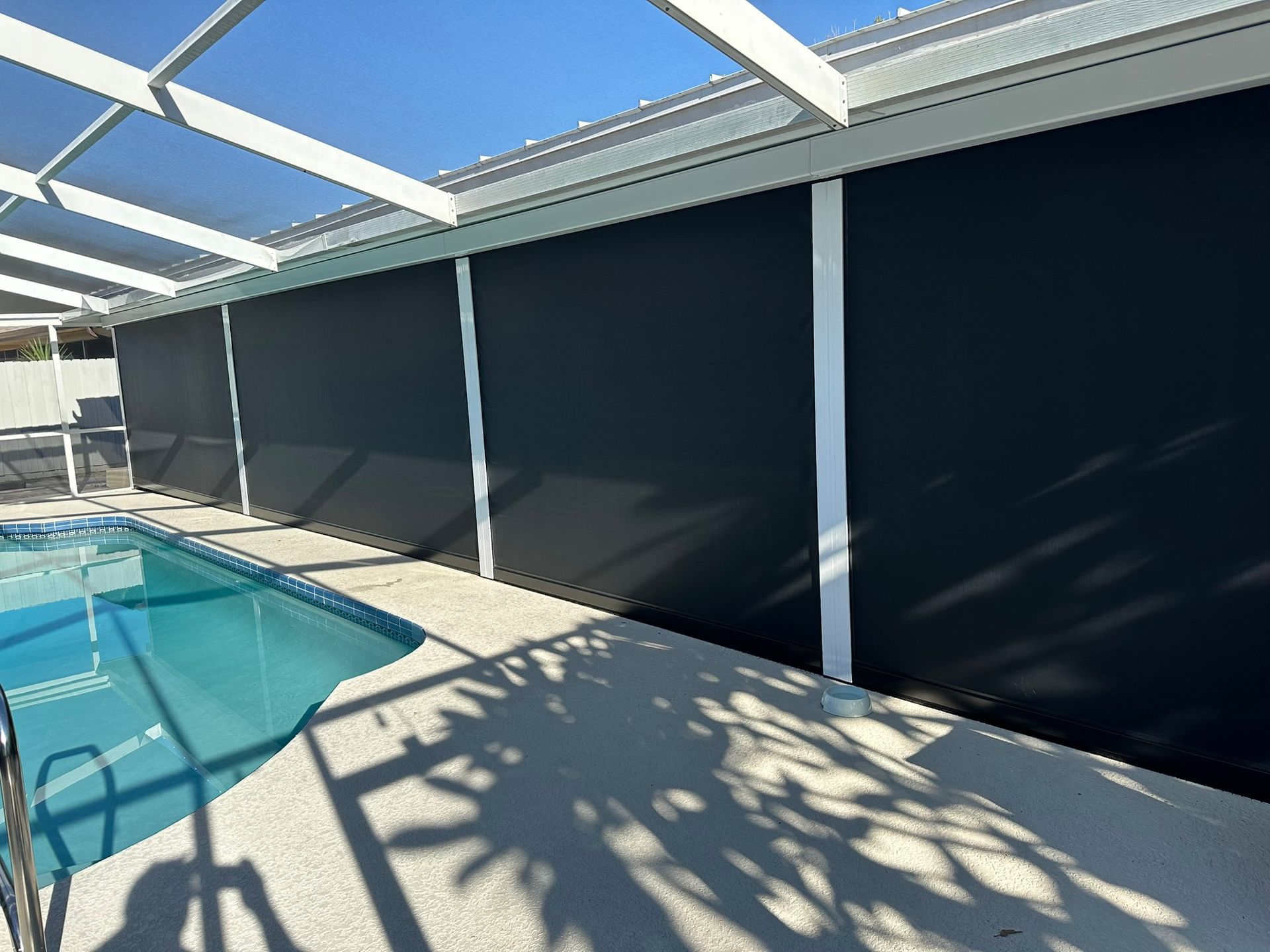 Screened poolside patio with black privacy shades and sunlight shadows beside a swimming pool