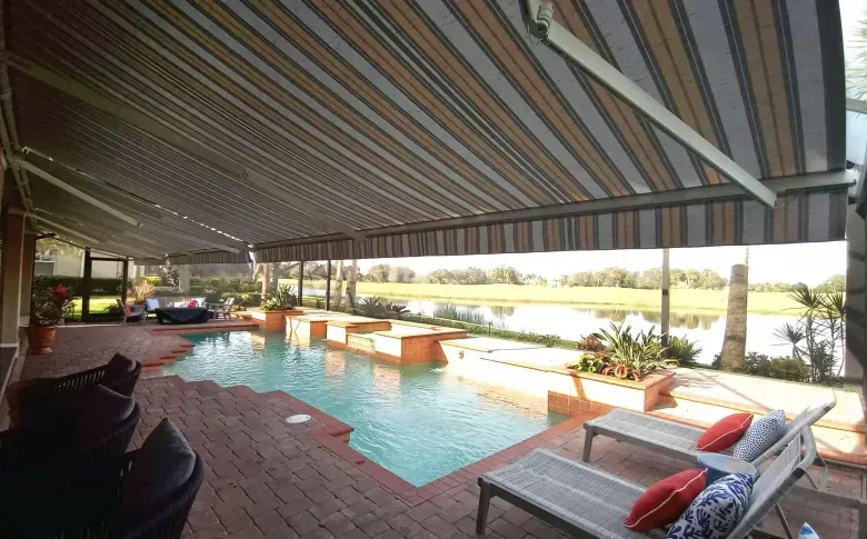 Covered patio with a pool, lounge chairs, and striped awning overlooking a sunny yard
