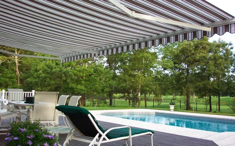 Patio with striped awning, white lounge chairs, and a swimming pool overlooking a green backyard