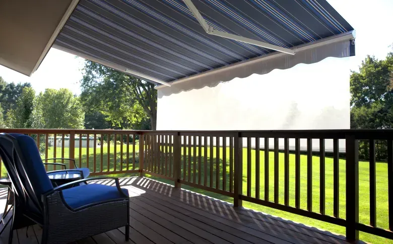 Covered wooden deck with blue chairs, railing, and a striped awning overlooking a green lawn.