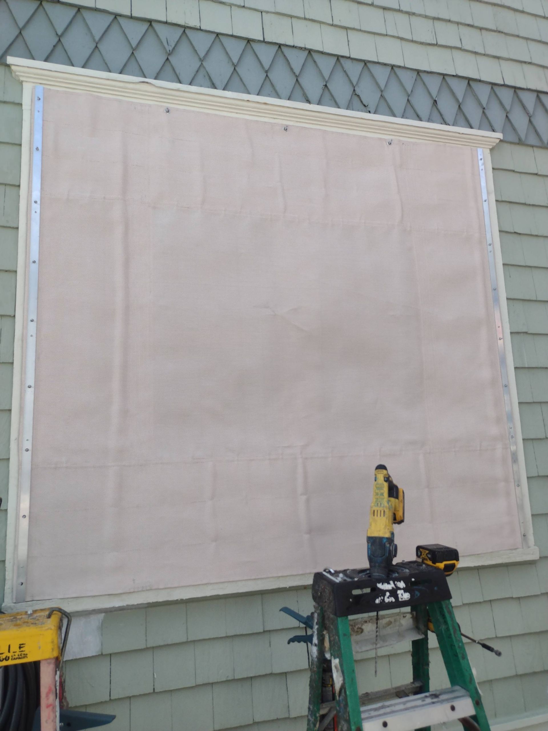 Large white fabric panel stretched on a wall, with a cordless drill on a small work platform in front.