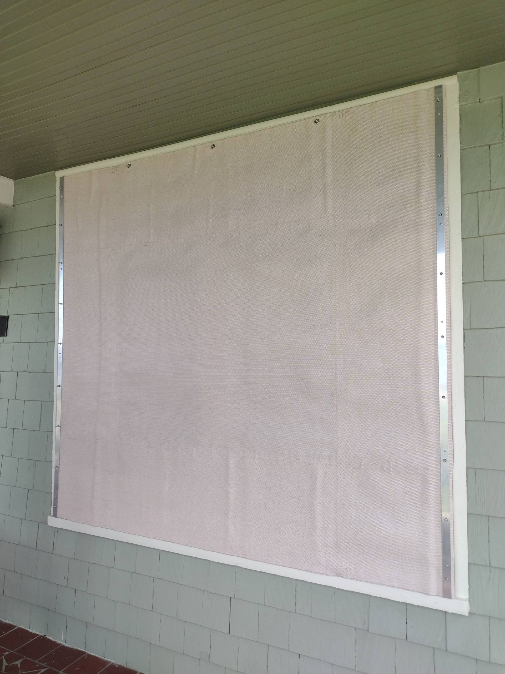White roller shade covering a large window on a light gray brick wall under a porch roof