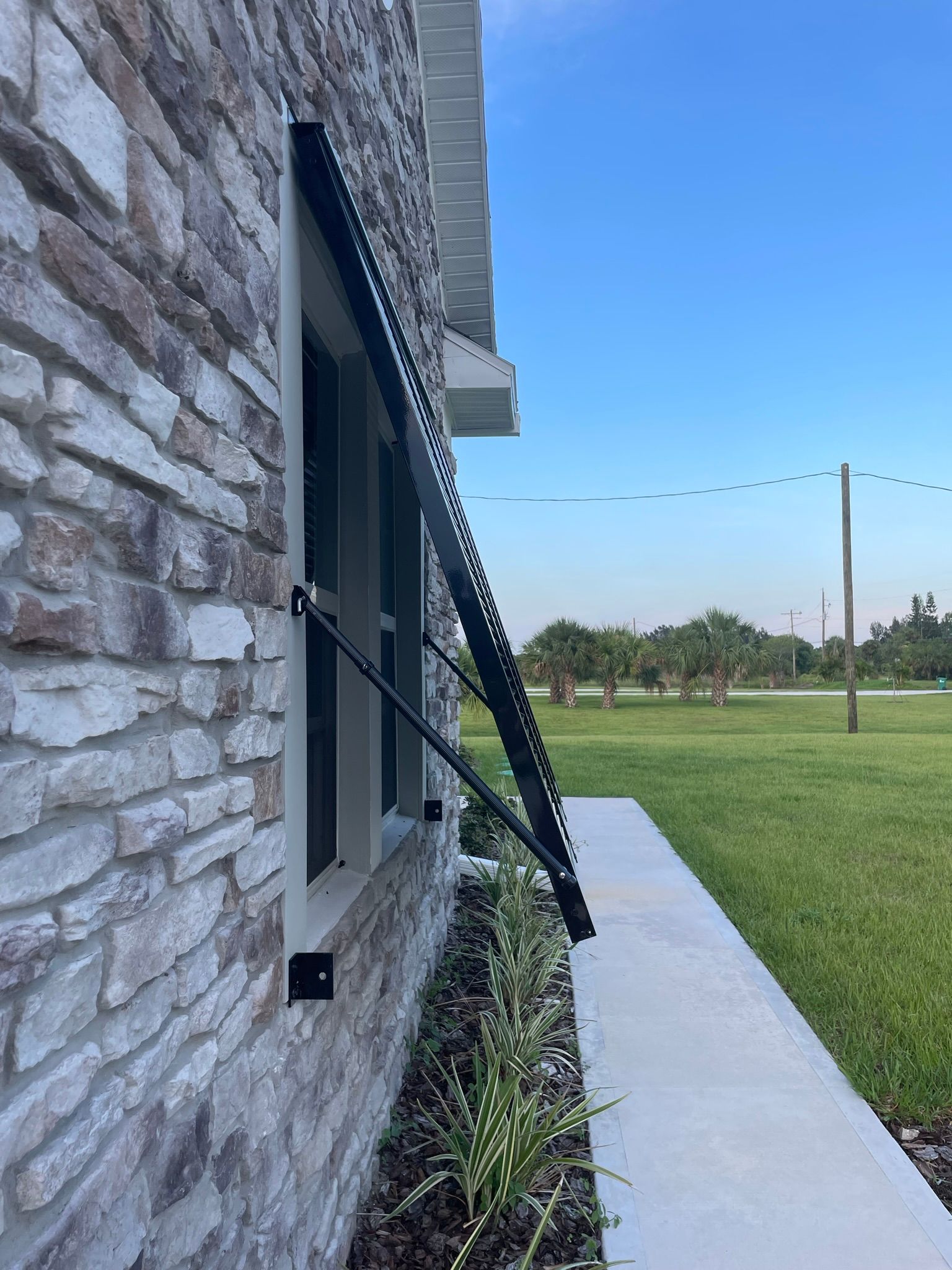 Stone house side view with a black window awning, sidewalk, and grassy field under a blue sky