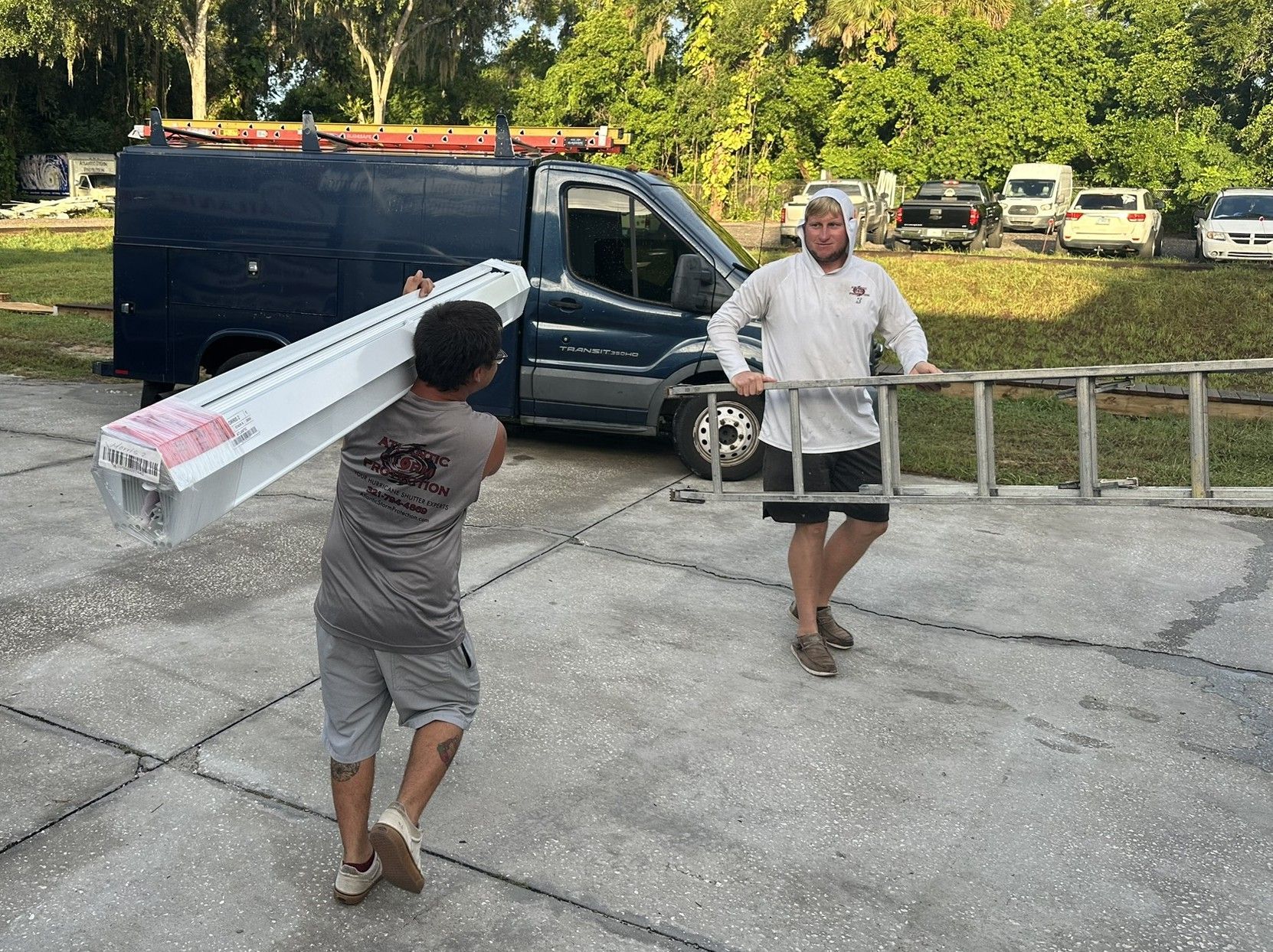 Two people carry a long white package near a black van in a driveway, with a ladder and trees nearby.