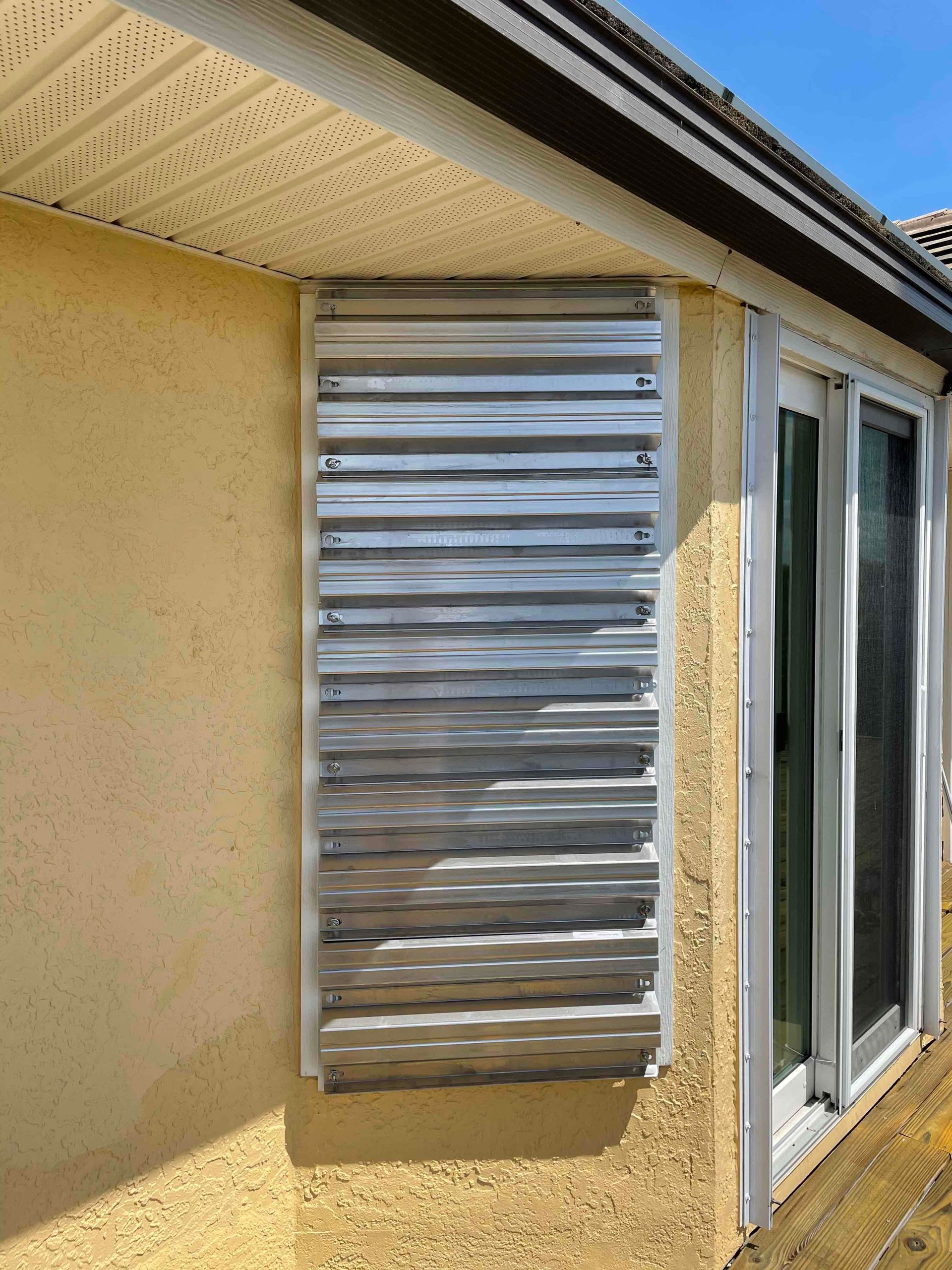 Silver exterior vent cover mounted on a tan stucco wall beside a sliding glass door