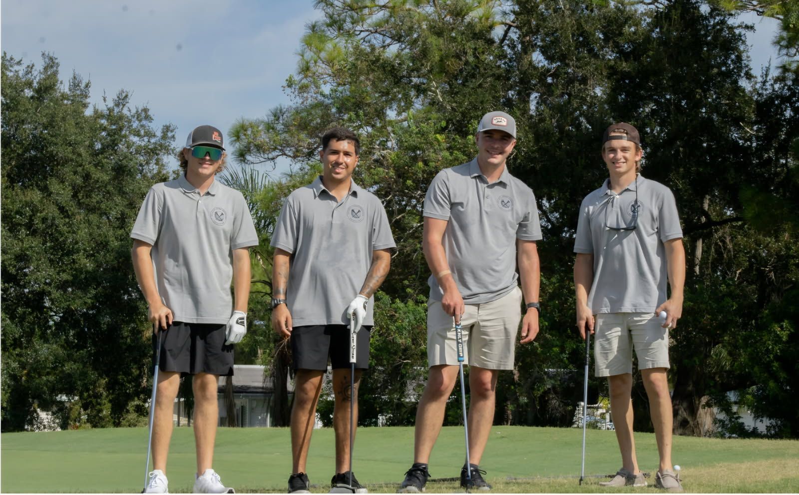 Four golfers in gray shirts and shorts standing on a golf course holding clubs