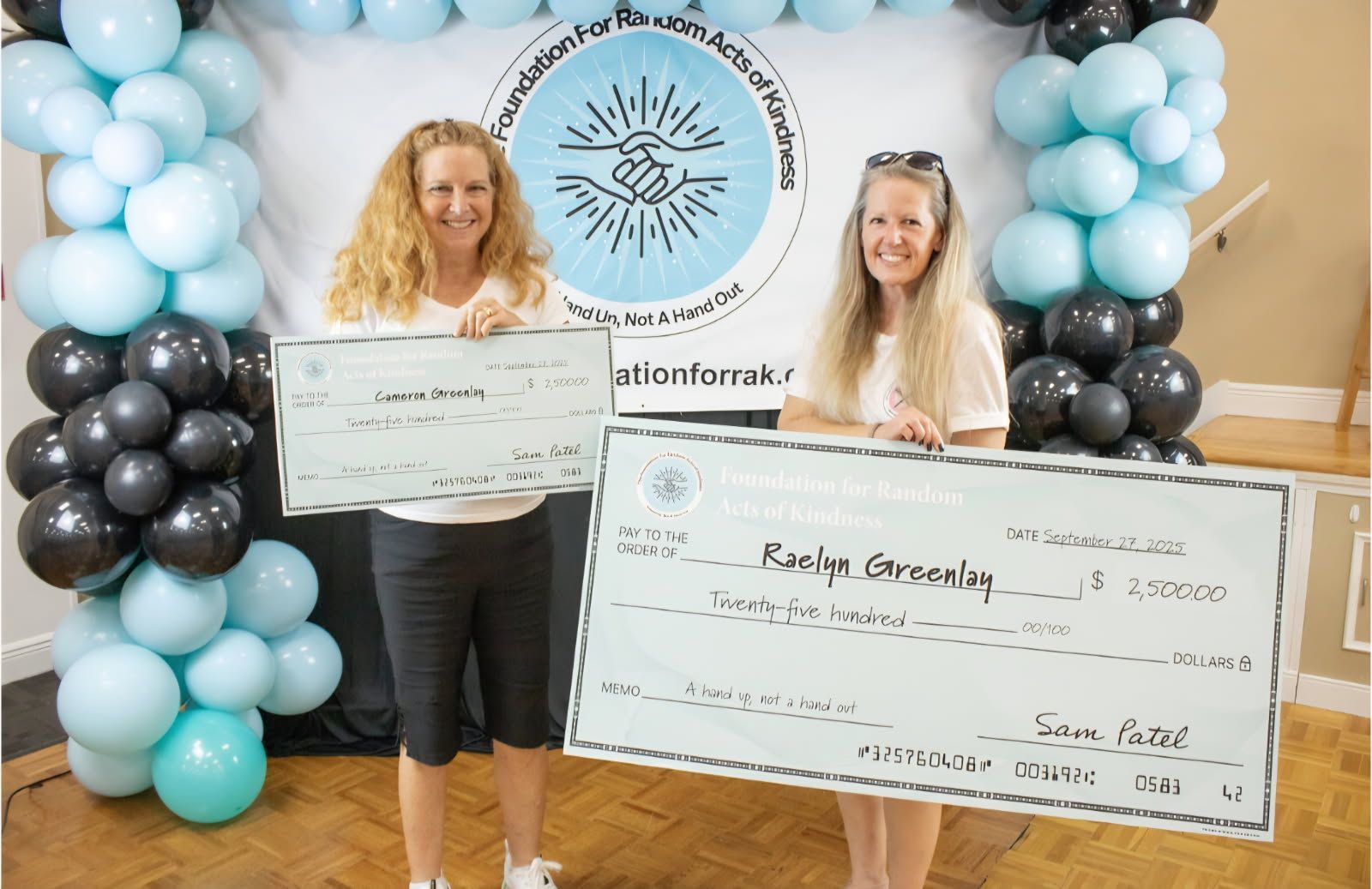 Two women hold oversized checks at a fundraising event with black, white, and blue balloon decorations.