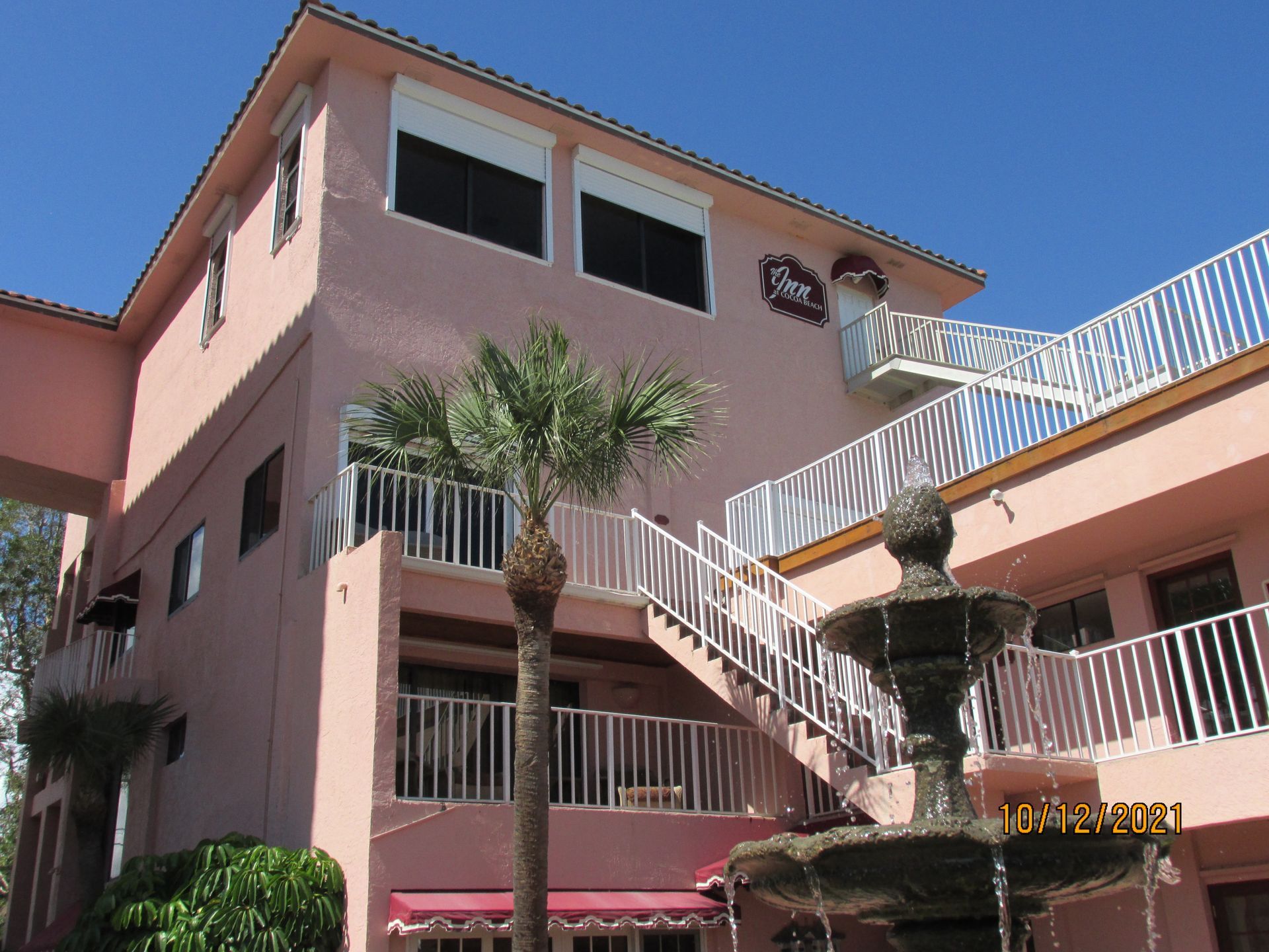 Pink stucco building with white railings, palm trees, and a staircase under a blue sky