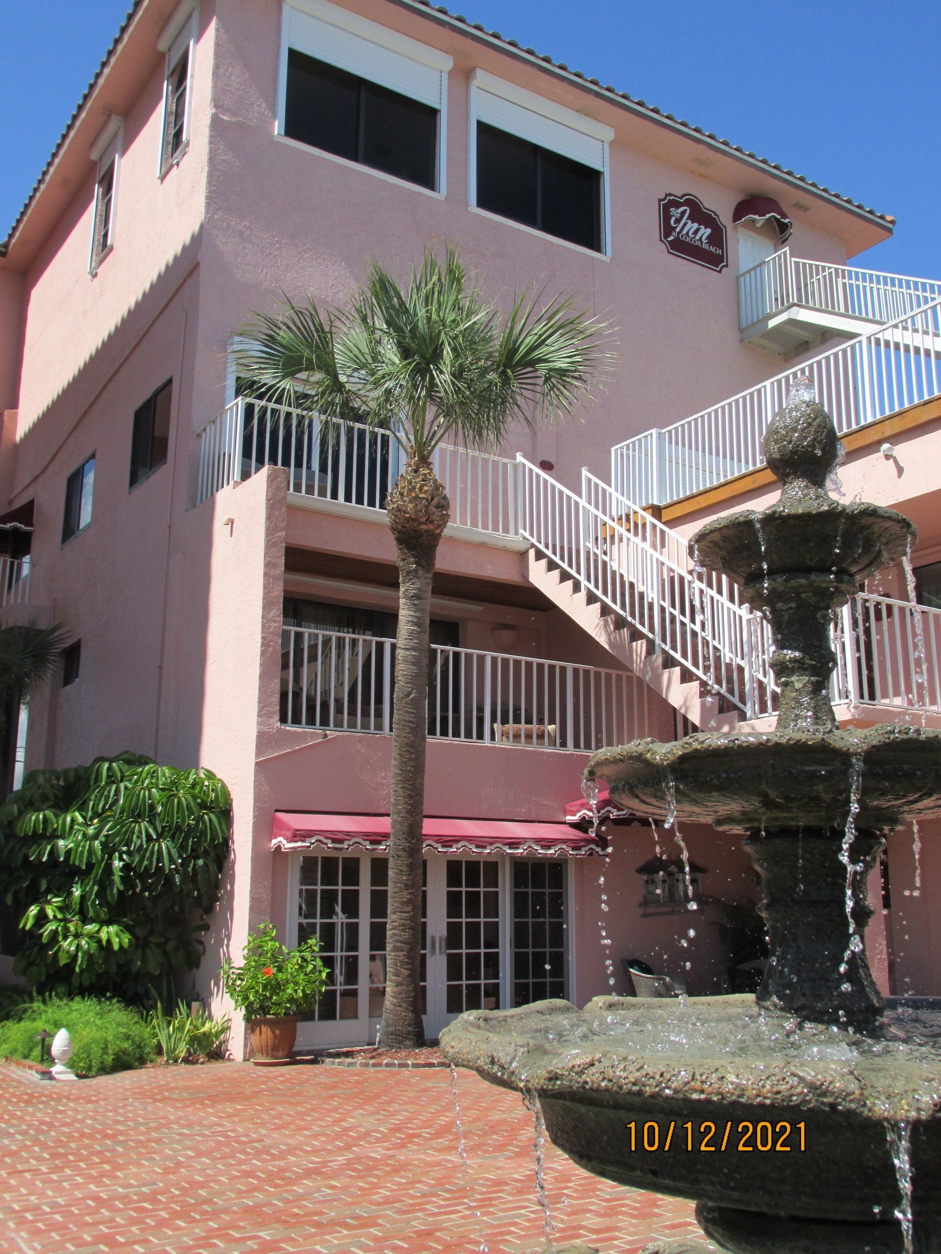 Pink stucco building with outdoor staircase, palm trees, and a fountain courtyard under a blue sky